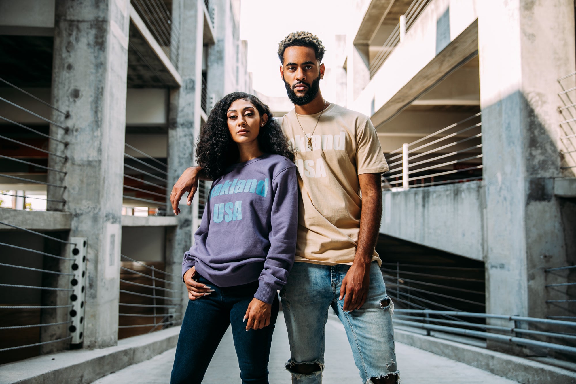 Guy and girl in parking garage structure, guy with tan tee with text Oakland USA, girl in purple crew with blue letters, same design.