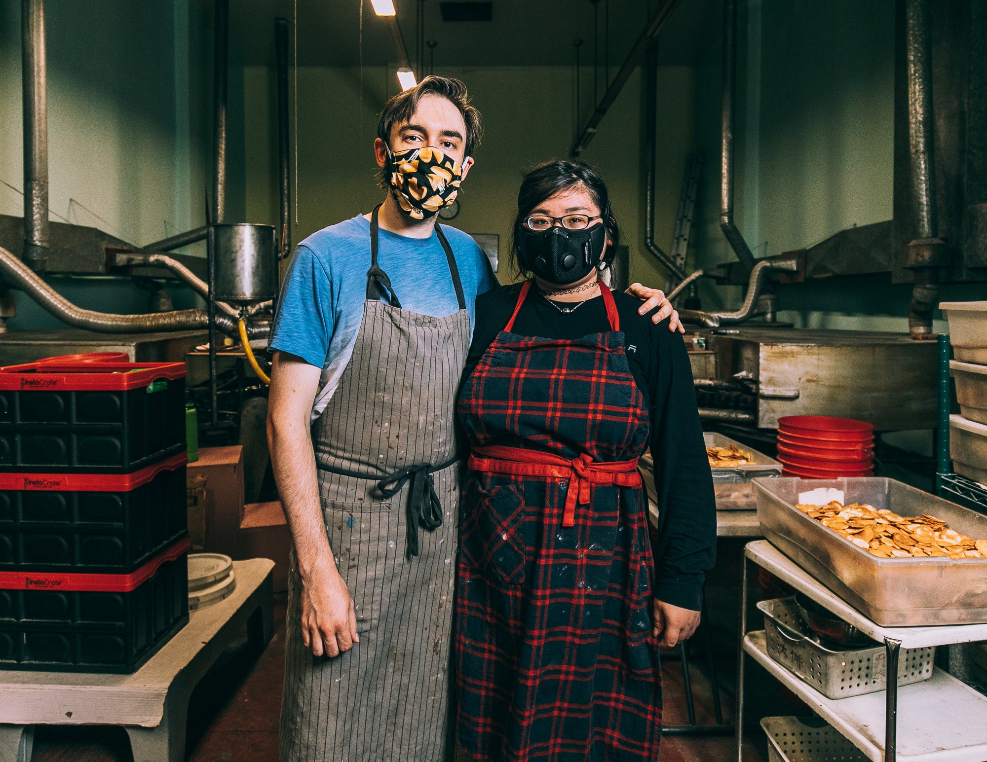 Alicia and Alex inside the fortune cookie factory, Alex has his arm around her shoulders.