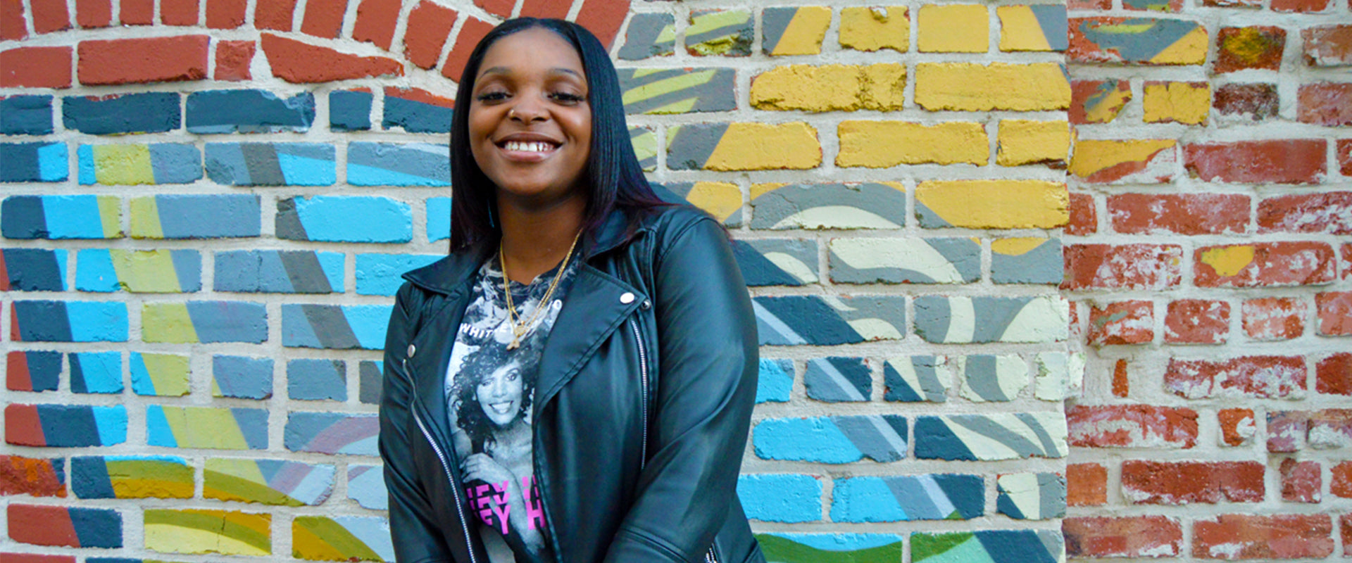 Amanni, smiling wearing jacket outdoors against a brick wall with mural.