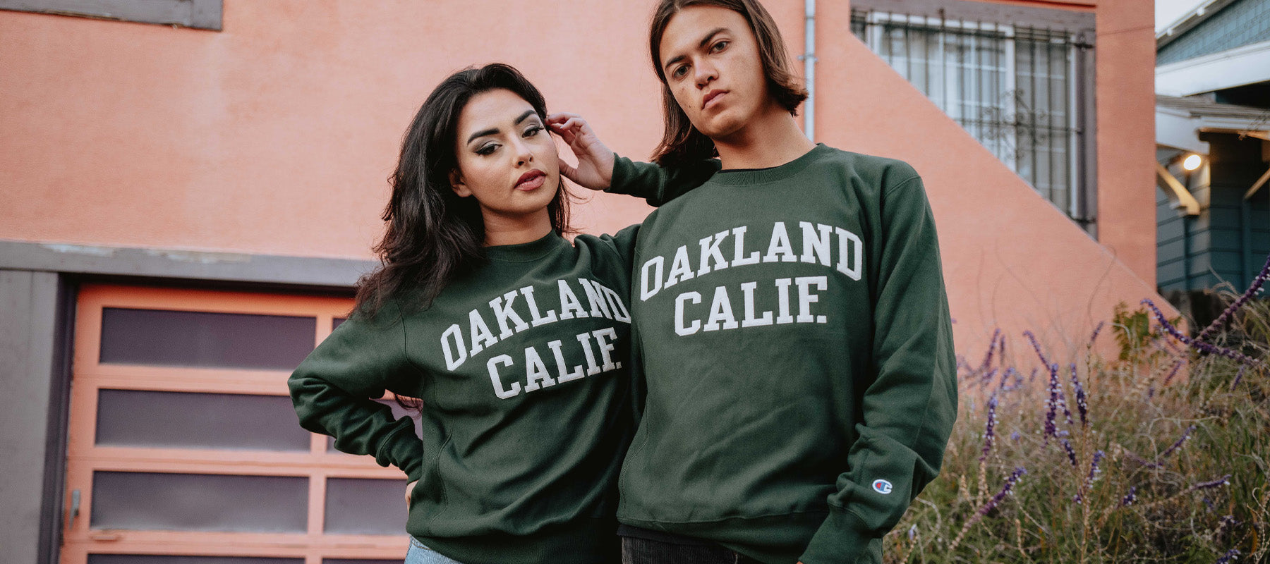 Woman and man outdoors in Oakland wearing a forest green Champion sweatshirts with OAKLAND CALIF. applique wordmarks.