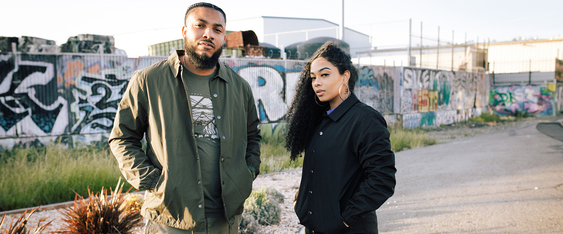 Man wearing an olive coaches jacket and women wearing a black coaches jacket in front of a wall on dirt path.