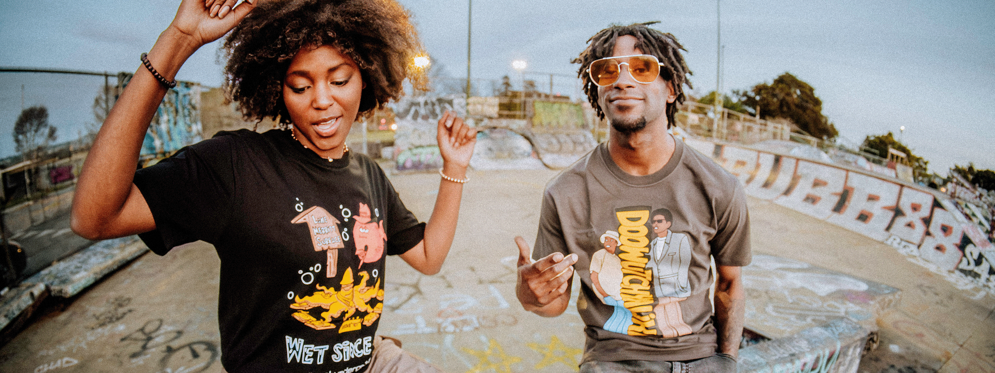 Two people dancing at a graffiti-covered skate park. They wear graphic t-shirts and have joyful expressions. The woman has curly hair and the man wears sunglasses. Skaters are visible in the background.