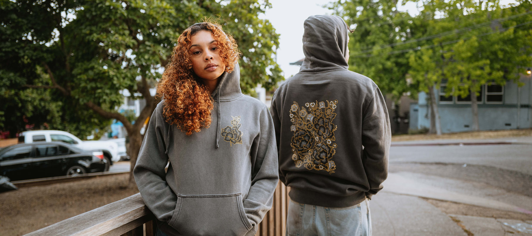 Man and Women on Oakland street in a black pigment-dyed black hoodie with bouquet flower design by artist Jet Martinez on his back.