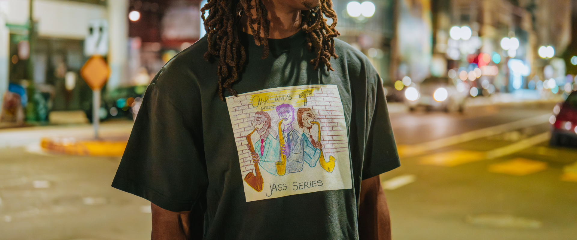 A man with long dreadlocks stands on a city street at night, wearing a dark T-shirt featuring colorful, hand-drawn art of people sitting and the text “The Projects Ass Series.” Blurred lights glow behind him.