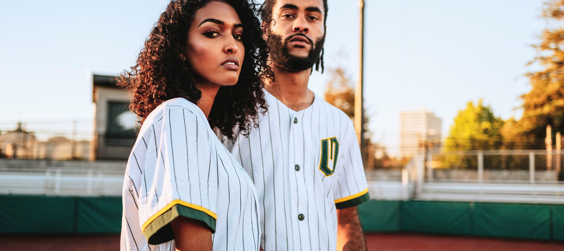 Man and woman on a baseball field in green striped white baseball jerseys with green and gold O for Oakland appliques on the chests.