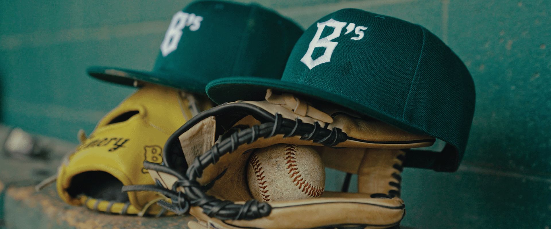 Two green baseball caps with "B's" logo rest on yellow baseball gloves and a baseball against a green wall. The caps and gloves are arranged neatly, suggesting a sports theme.