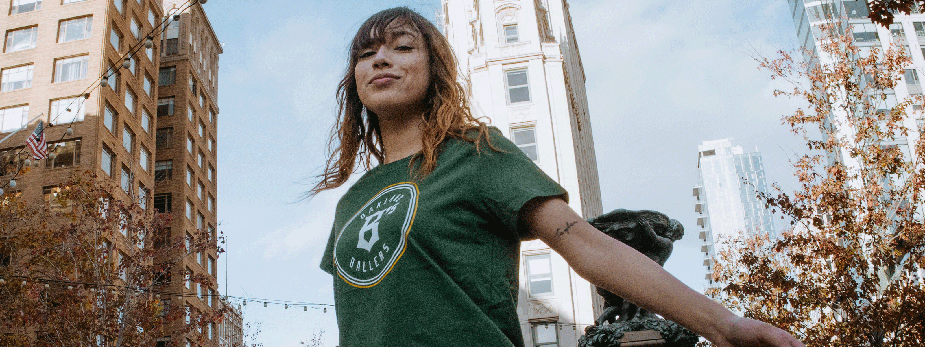 A woman in a green T-shirt stands in a city plaza, surrounded by tall buildings and autumn trees. She smiles confidently with her arms outstretched under a clear blue sky.