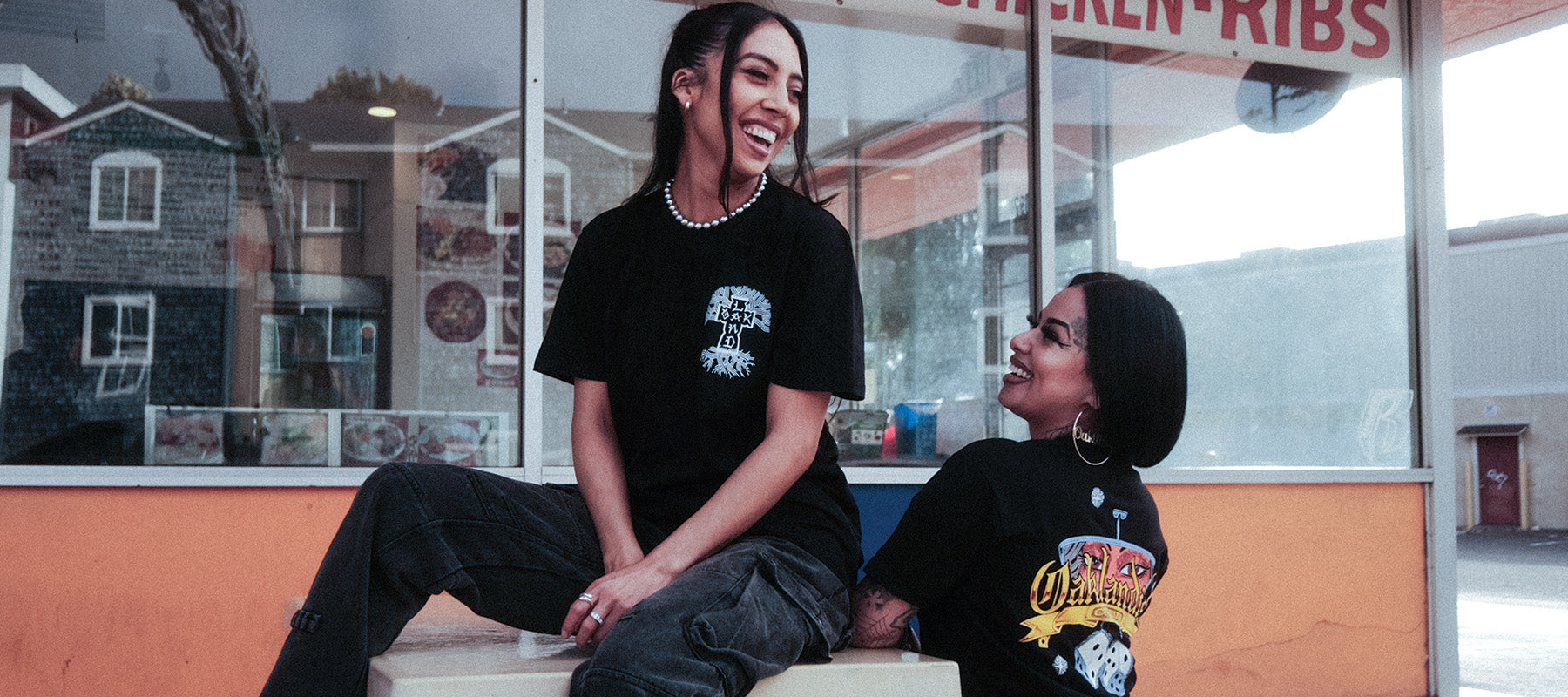 Two women seated outdoors in front of a restaurant, smiling and wearing black tees with left chest and back print.