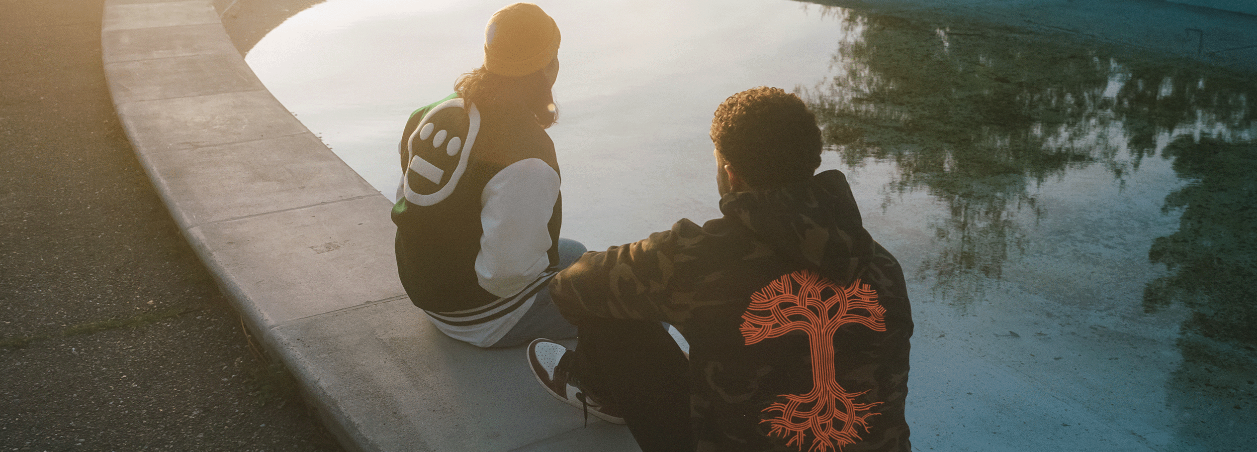 Two people sit at the edge of a pool, backs to the camera. One wears a varsity jacket with a white symbol, the other a camo jacket with an orange tree design. The scene is softly lit by sunset.