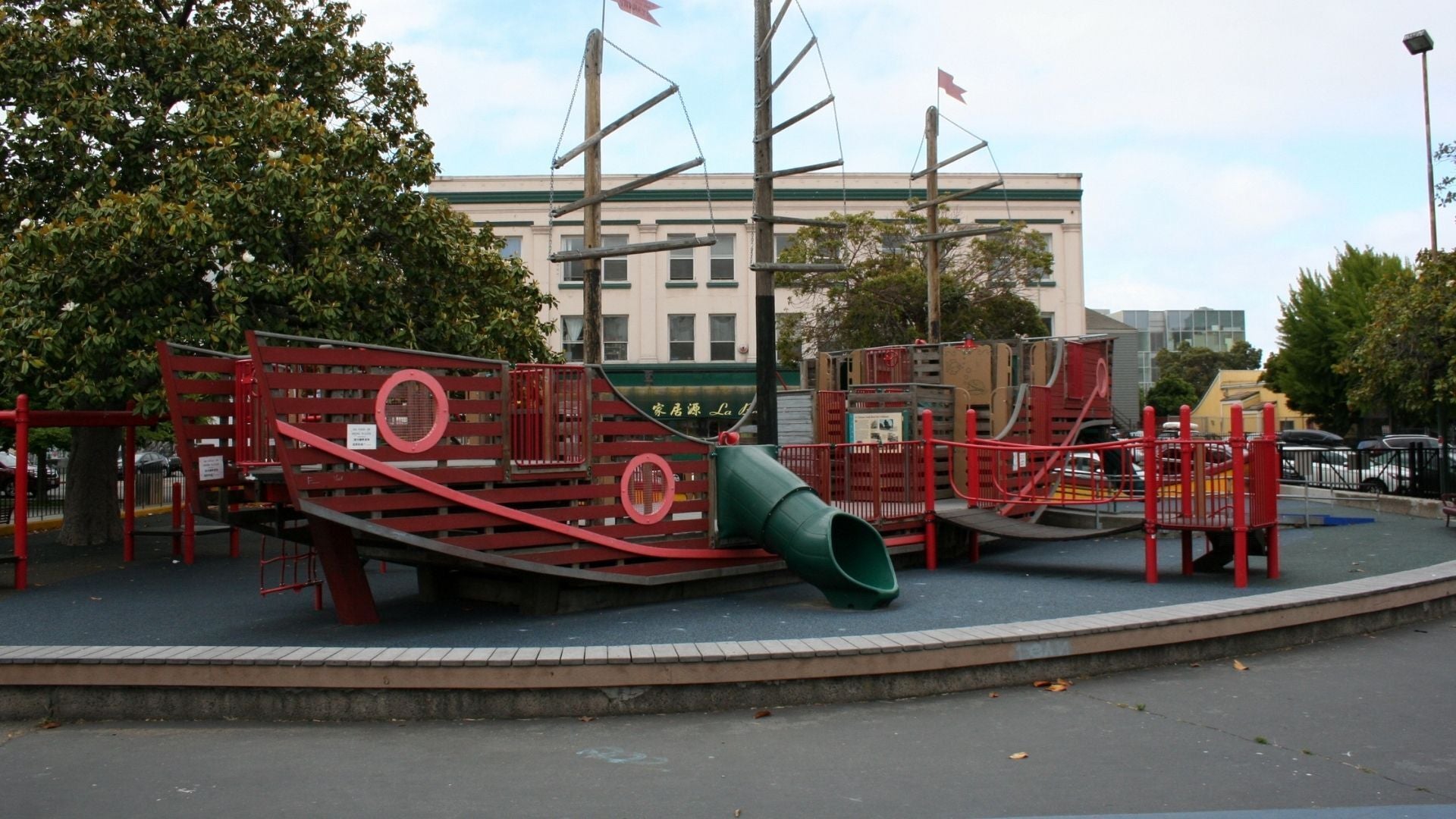 A playground shaped like a red wooden pirate ship with a green slide, climbing areas, and nautical details, set on rubber flooring with trees and a building in the background.
