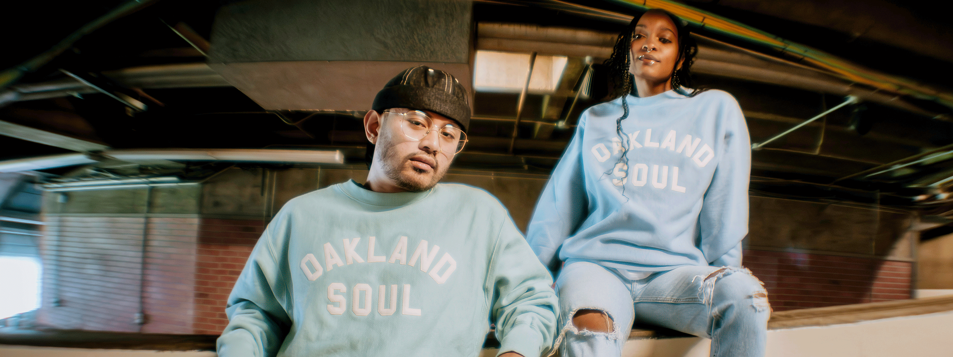 Four soccer players stand side by side on a stadium balcony overlooking a field. Two wear black geometic jerseys, and two wear white with green, black, and yellow accents. Each holds a soccer ball.