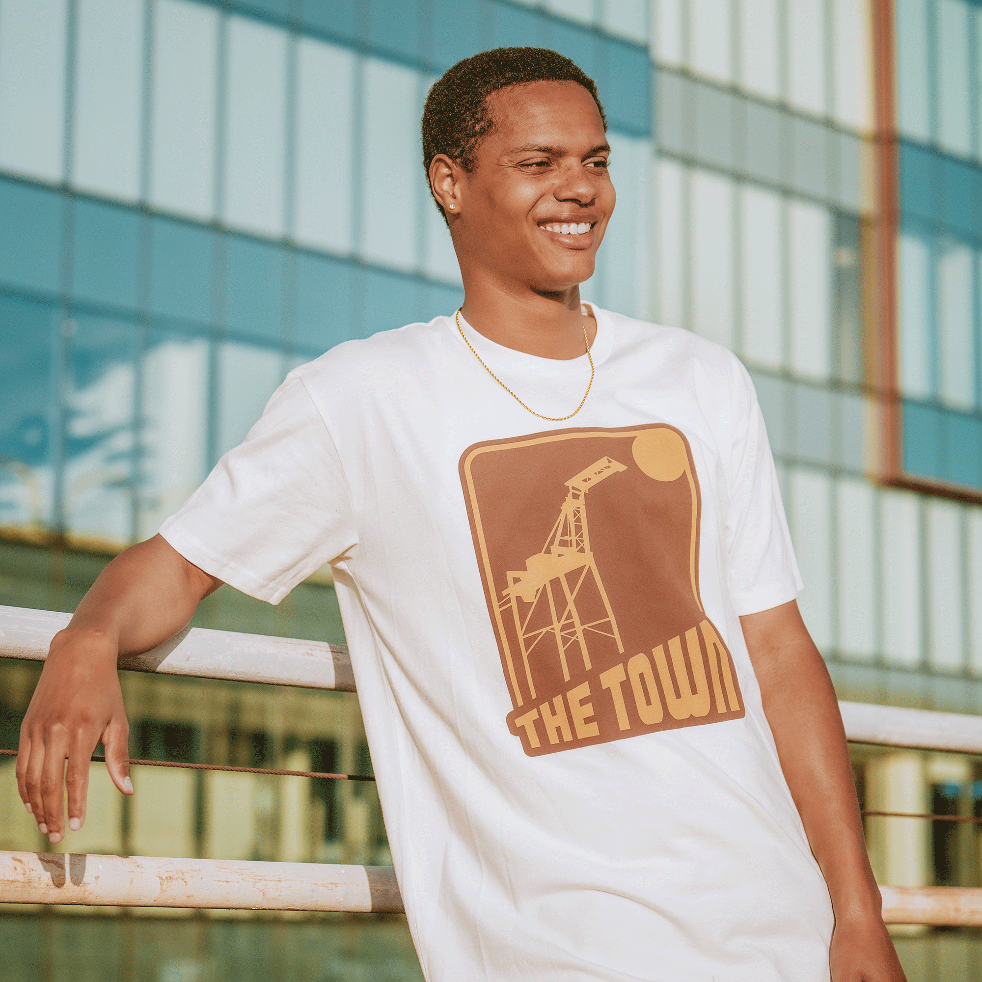 A smiling young man stands outdoors by a glass building, leaning on a metal rail. He wears a GSW 70's Trading Card Tee paired with a gold necklace.
