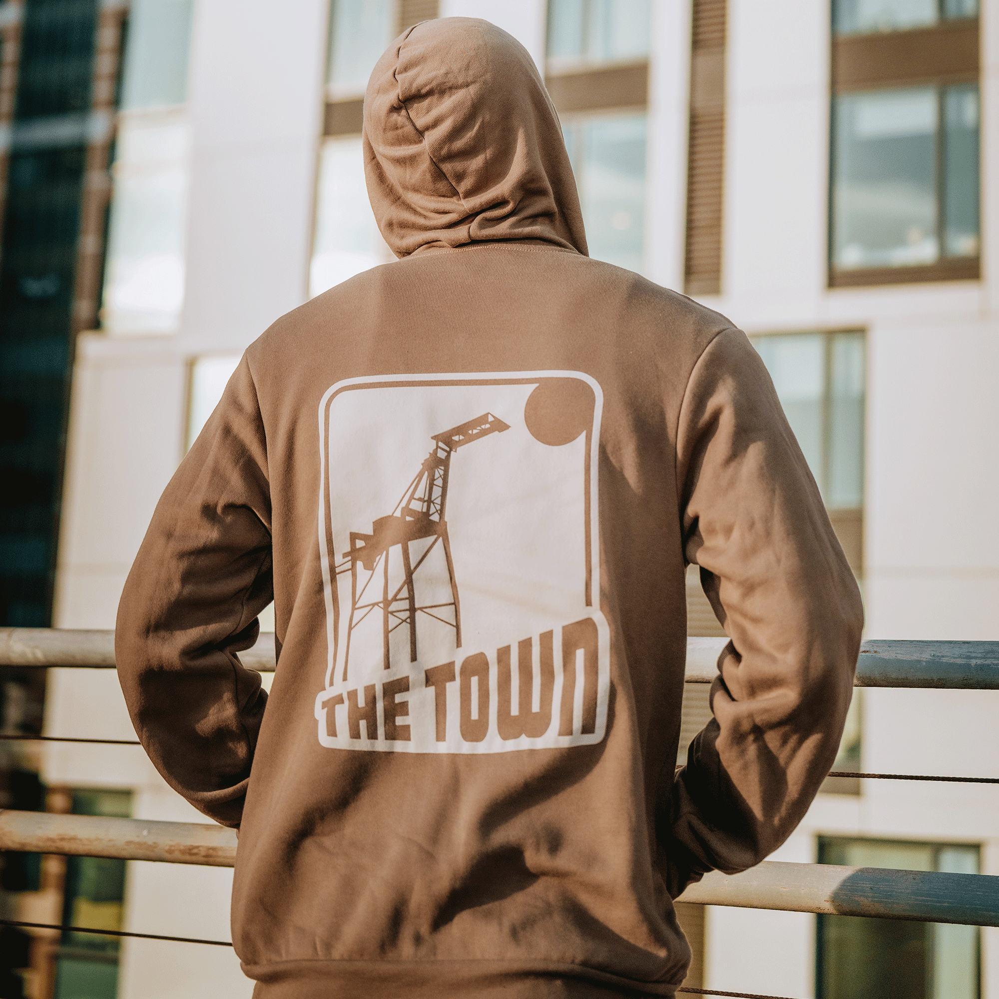 A person in a GSW 70’s Trading Card Zip Hoodie stands with their back to the camera. The graphic features a crane, sun, and “THE TOWN,” set outdoors by a modern building with tall windows and a metal railing.