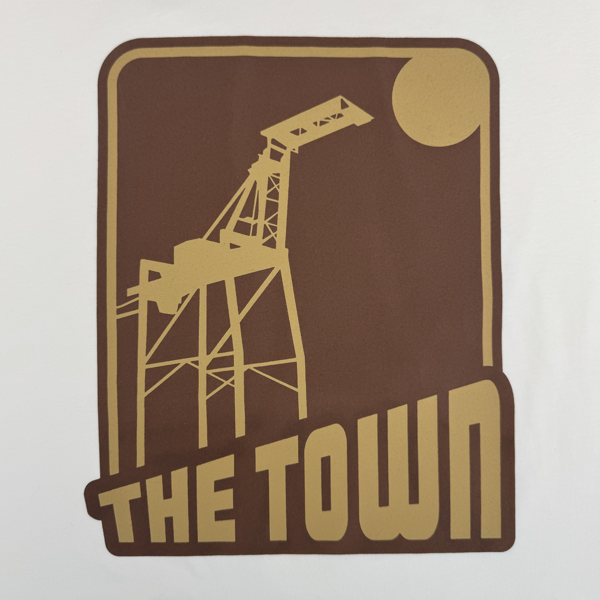 A brown and gold graphic on the GSW 70’s Trading Card Tee shows a large industrial crane with a sun or moon behind it, plus bold gold “THE TOWN” text, inspired by Oaklandish and the Town Collection in retro minimalist style.