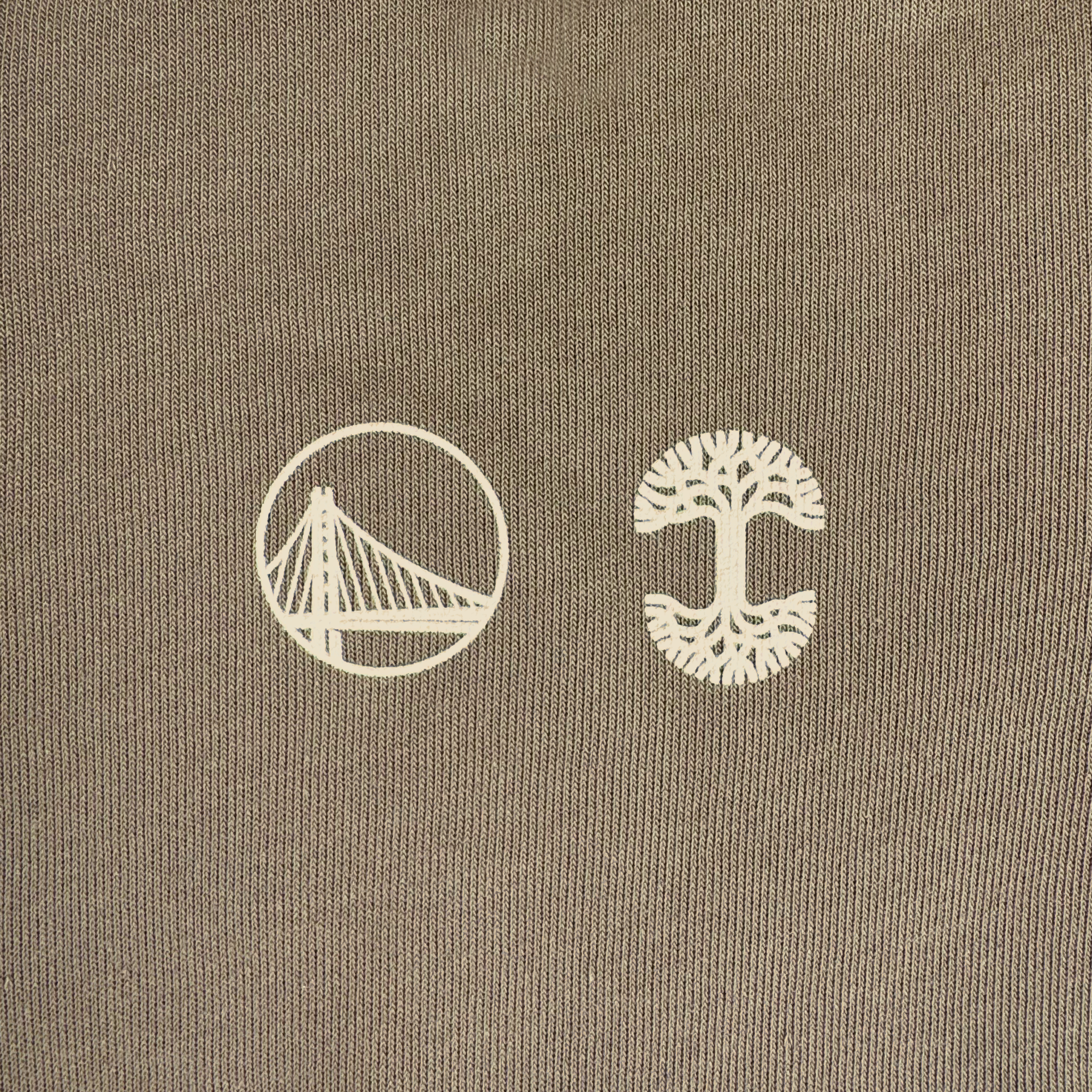 A close-up of brown fabric from the GSW 70’s Trading Card Zip Hoodie displays two white graphics: a cable-stayed bridge in a circle on the left and a stylized tree on the right, reflecting Oaklandish’s signature minimalist aesthetic.