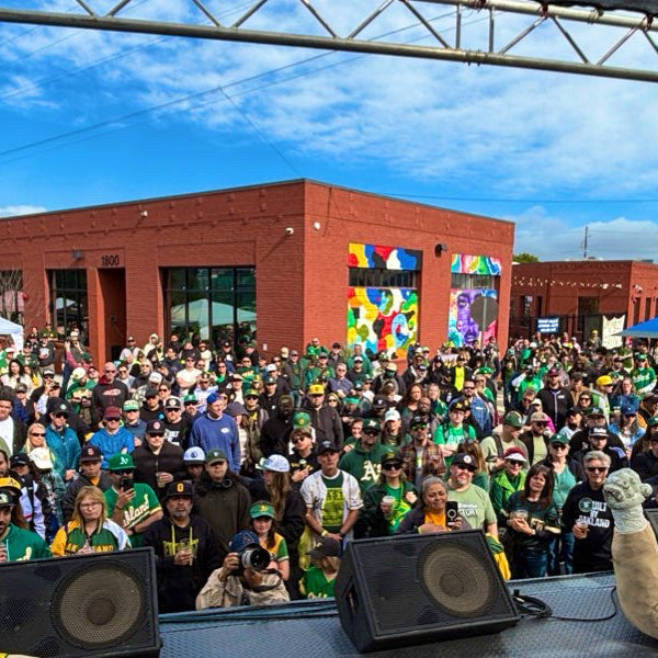 A lively crowd in green and yellow gathers outside a mural-covered red brick building under blue skies at Oakland68s’ Fans Fest 2026 — Community Donation, captured from the stage with speakers visible, showing community spirit and excitement.