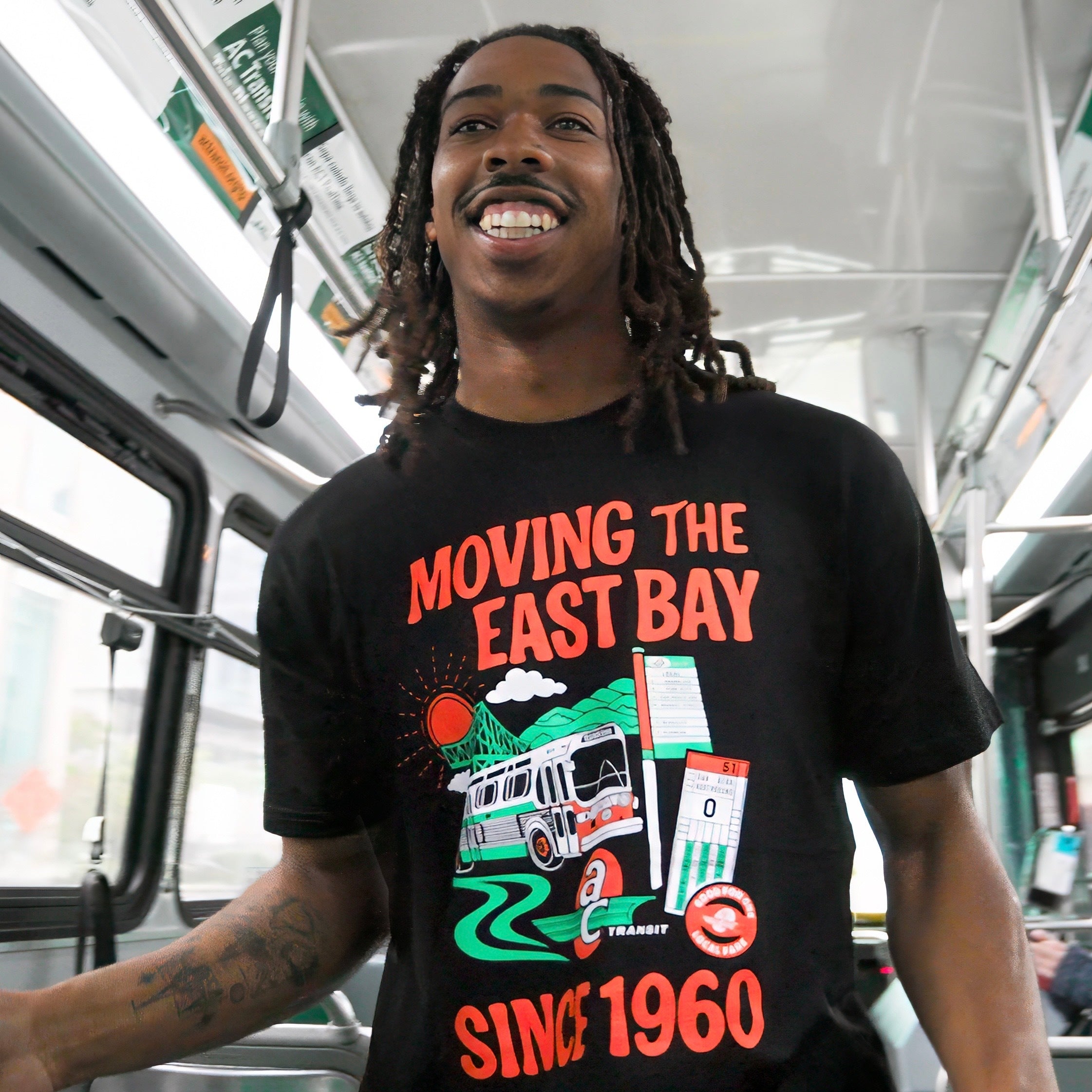 A smiling young man with dreadlocks stands on an AC Transit bus, wearing the Oaklandish "AC Transit Moves The East Bay Tee" featuring a vibrant bus, sun, and route signs graphic. Bright light fills the space.