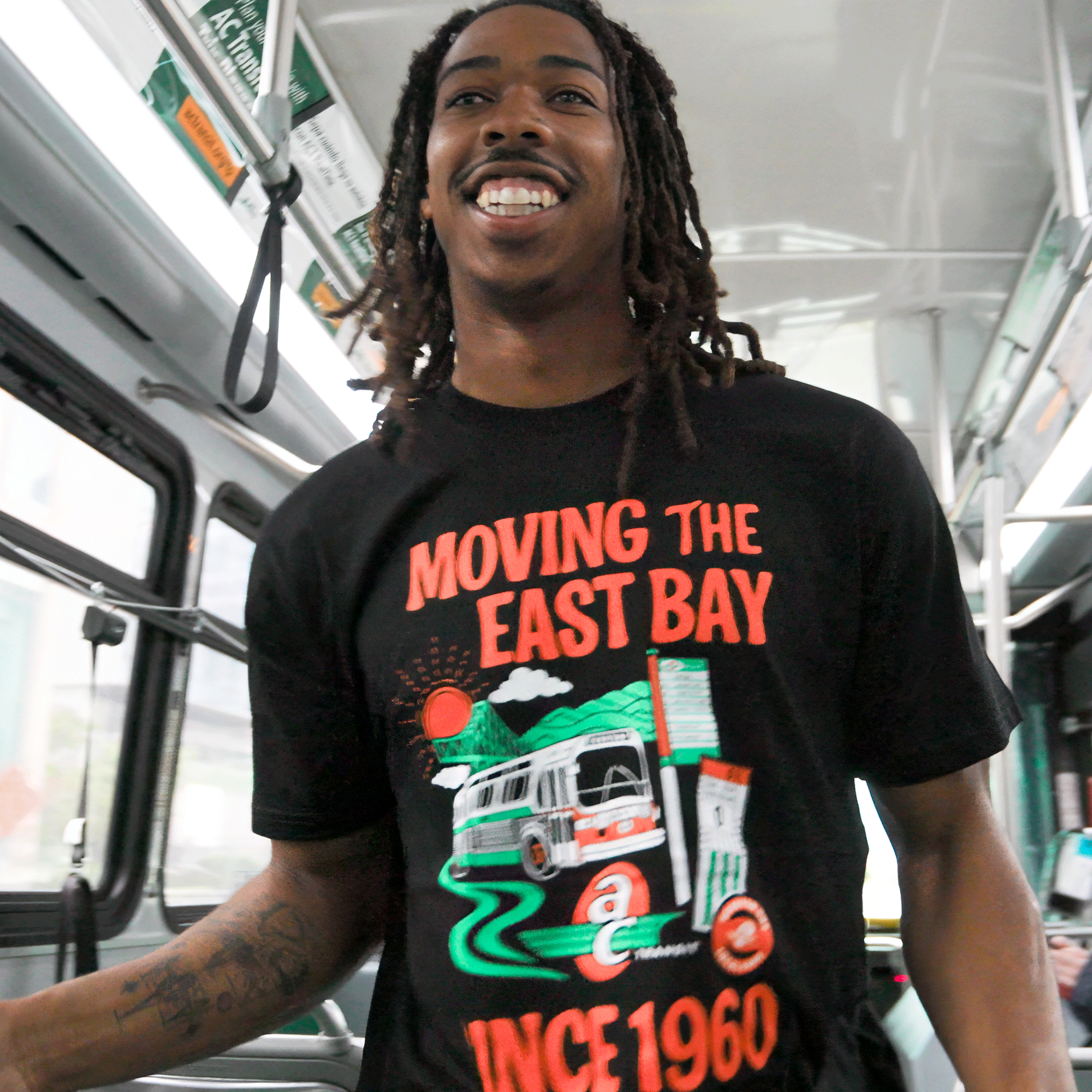 A smiling young man with dreadlocks stands on a bus wearing the Oaklandish AC Transit Moves The East Bay Tee. The T-shirt features a colorful graphic and “MOVING THE EAST BAY SINCE 1960” slogan; the AC Transit bus interior is visible behind him.