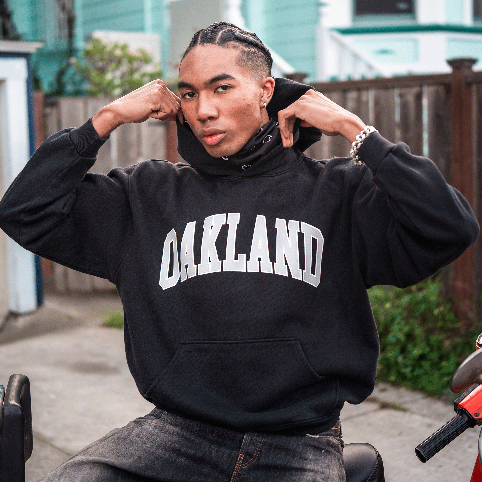 A young man with braided hair sits on a black bike outdoors, wearing the oversized Alma Mater Hoodie 2.0 by Oaklandish with bold "OAKLAND" lettering. He raises the hood confidently, gazing at the camera; pastel houses and a wooden fence are behind him.