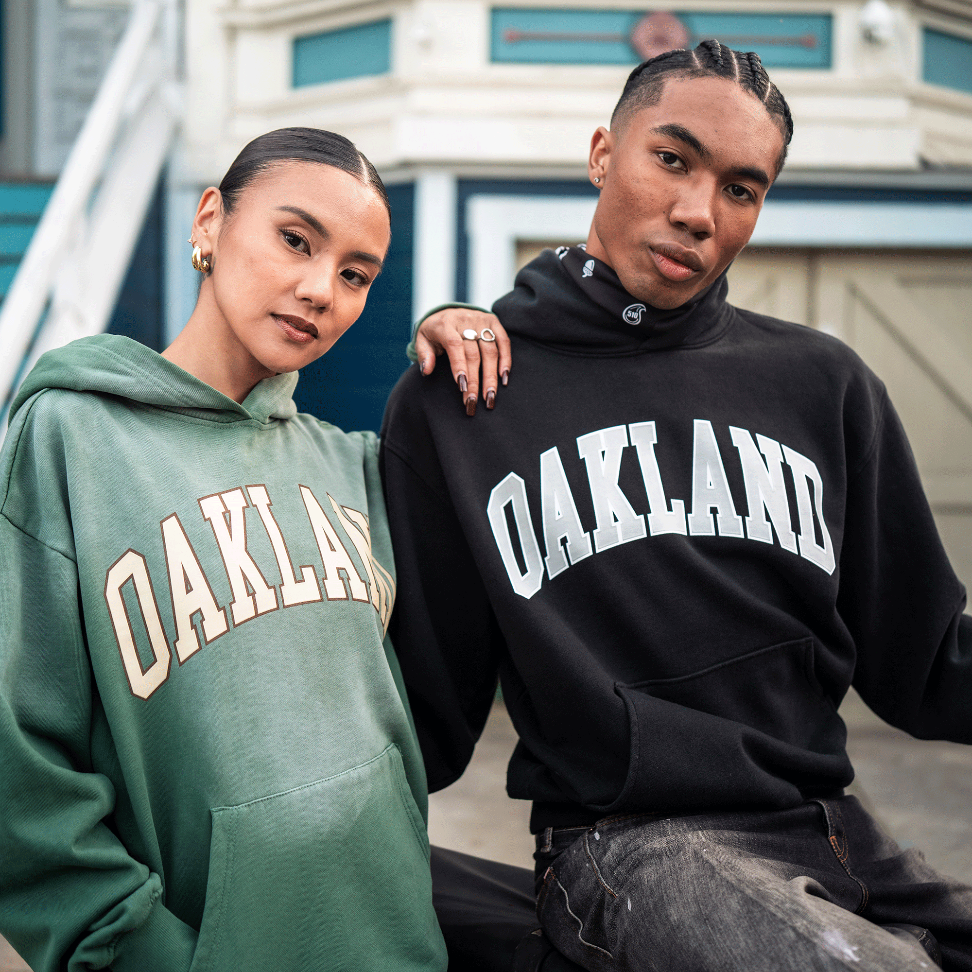 Two people sit side by side outdoors, each wearing an Alma Mater Hoodie 2.0 from Oaklandish—one in green men's pullover and the other in black oversized fleece. Both lean together, gazing confidently at the camera with a blurred building and stairs behind them.