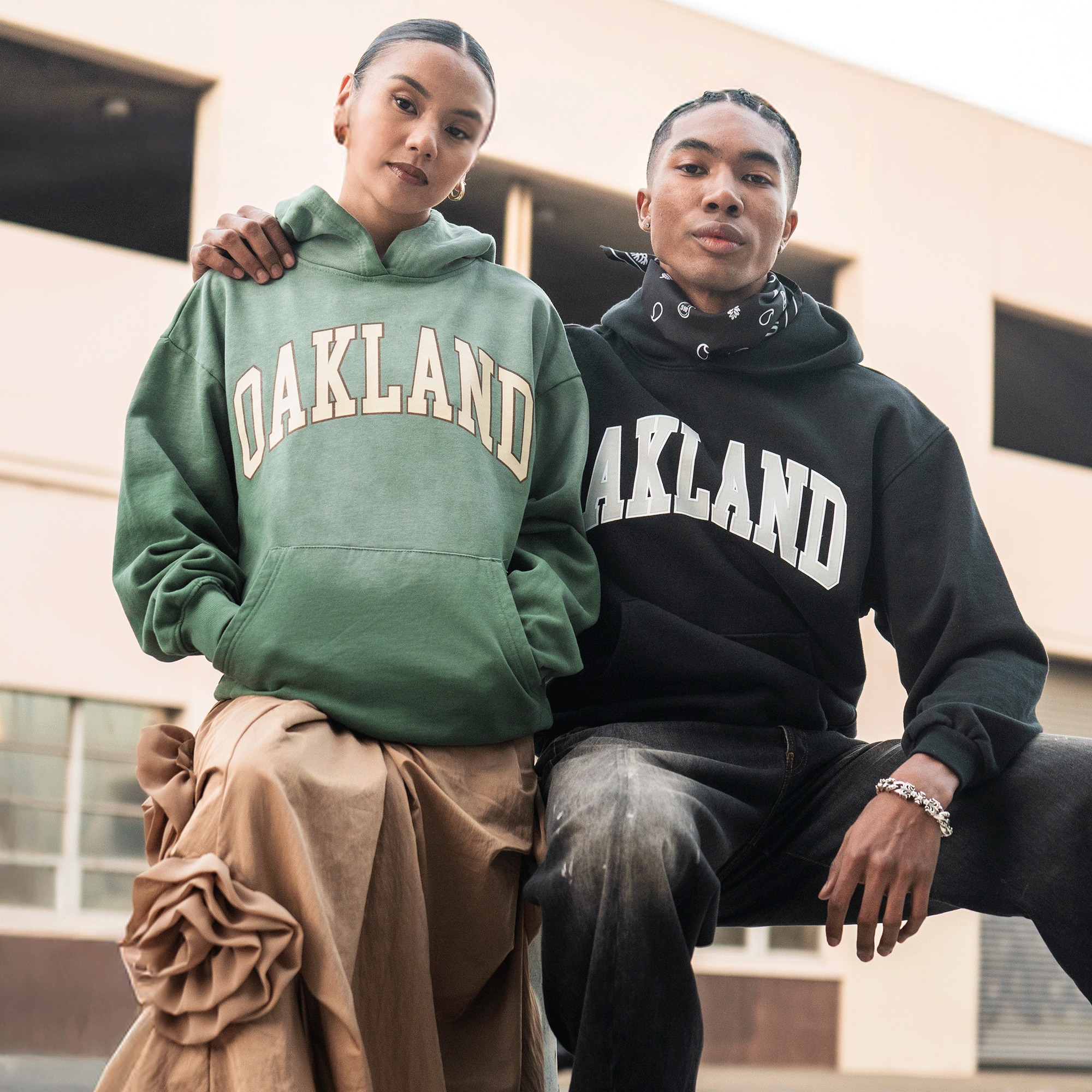 Two people pose confidently outdoors in Oaklandish Alma Mater Hoodie 2.0—one in green with a tan skirt, the other in a men's pullover—both crafted from 100% cotton, sitting close together by an industrial building.