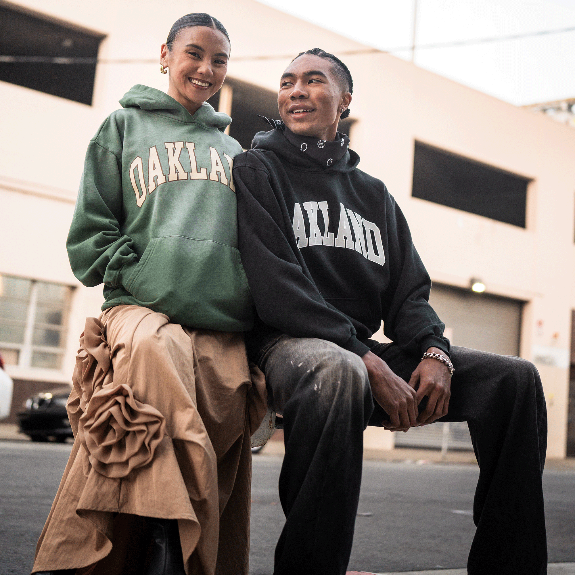 Two people smile outdoors in Oaklandish Alma Mater Hoodie 2.0 sweatshirts—one wears a green oversized fleece with a tan ruffled skirt, the other a black pullover with black jeans. Blurred city buildings and street scenes create the background.