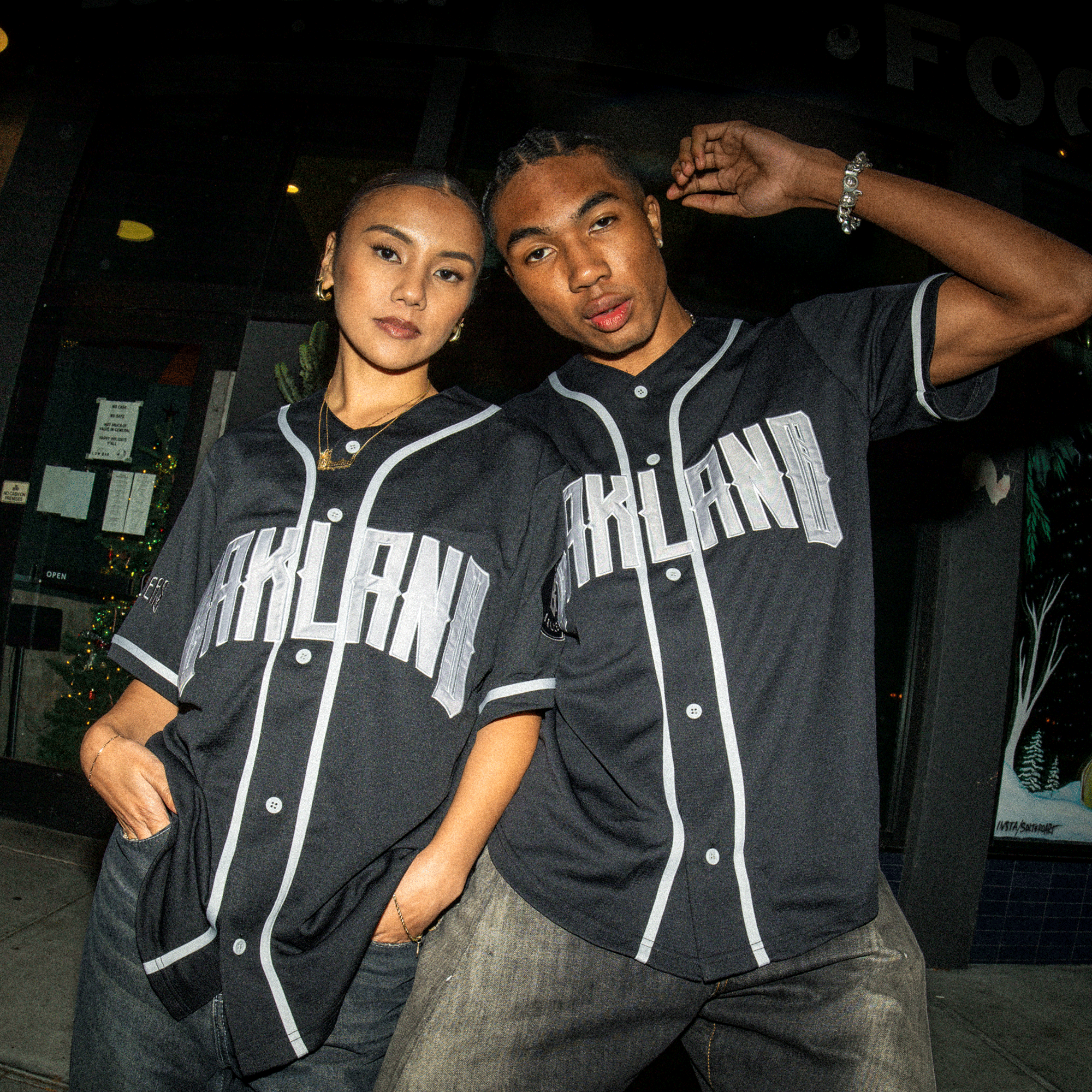 Two young adults pose confidently outside at night, both wearing matching black Oakland Ballers Button Up Jerseys with white trim. One has an arm over the other's shoulder as they stand before a glowing storefront. Brand: Oakland Ballers.