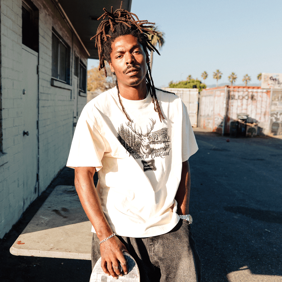A Black man with dreadlocks wears a cream-colored, heavy-weight cotton Oaklandish Buck Tee featuring a deer illustration, standing outdoors in an urban setting with his hand on a skateboard, wearing dark jeans and a silver watch.