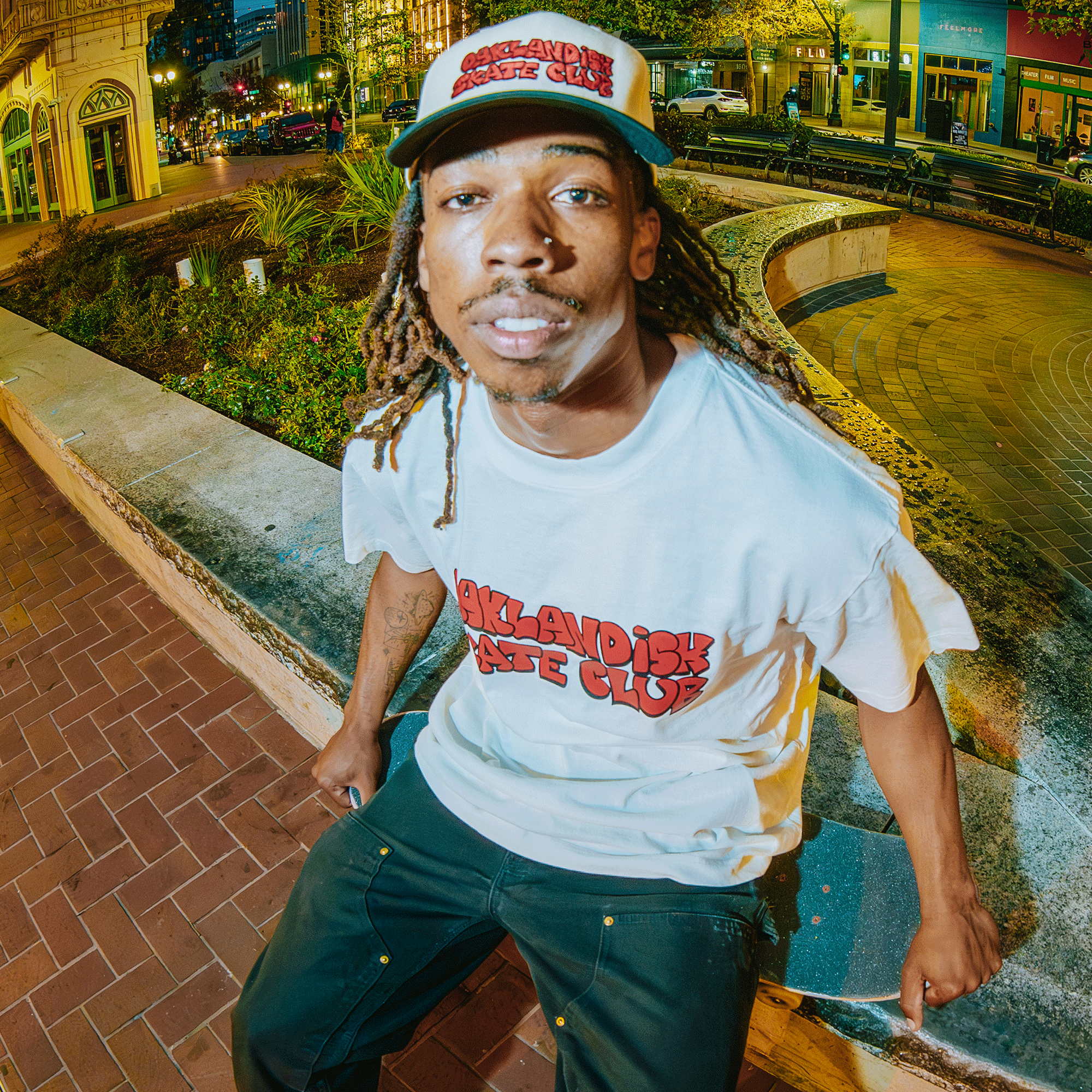 A man with long dreadlocks, a mustache, and beard sits on a curved concrete bench at night in the city, wearing an Oaklandish Skate Club 2-Tone Hat by Oaklandish and a white t-shirt, capturing skateboard culture under urban lights.