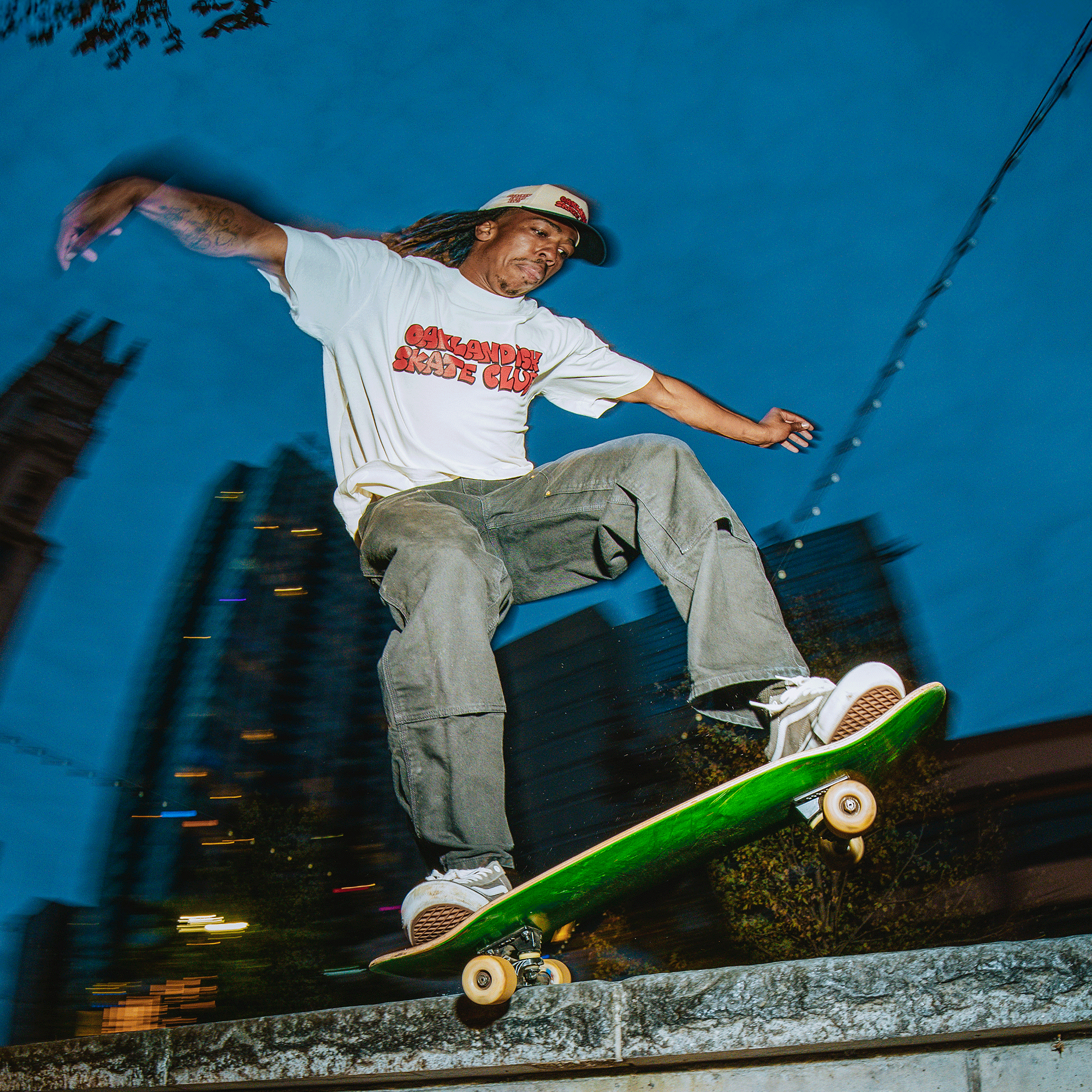 Wearing the Oaklandish Skate Club Tee and gray pants, a skateboarder grinds a ledge on a green board at dusk. The city skyline glows behind him, capturing Oaklandish style and true skateboard culture against the blue evening sky.