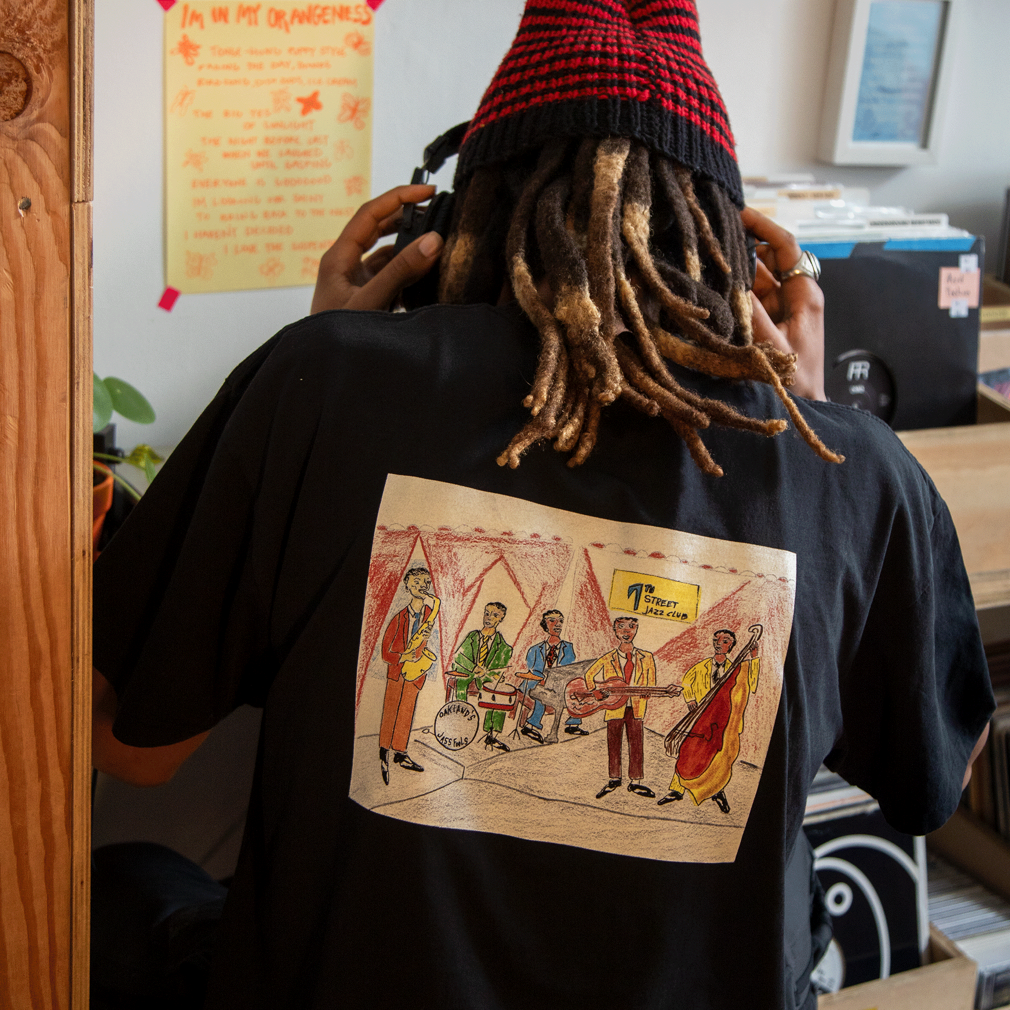Wearing the Oaklandish Ode to Jass Tee, a person with long, light brown dreadlocks stands among records and colorful posters. The classic fit 100% cotton shirt features a jazz band illustration. They also wear a black and red beanie and headphones.