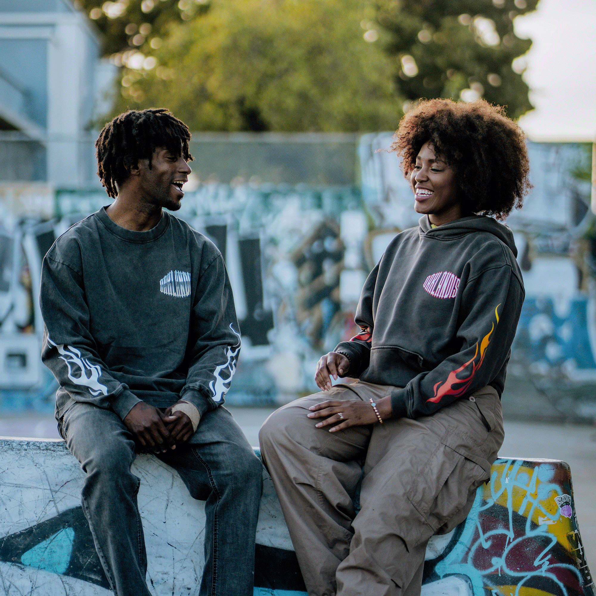 Two people in casual streetwear sit on a graffiti-covered ledge, smiling and engaged in conversation. One wears the Oaklandish Chopper Heavy LS Tee with vintage black flame patterns on the sleeves. The blurred background features street art and trees.