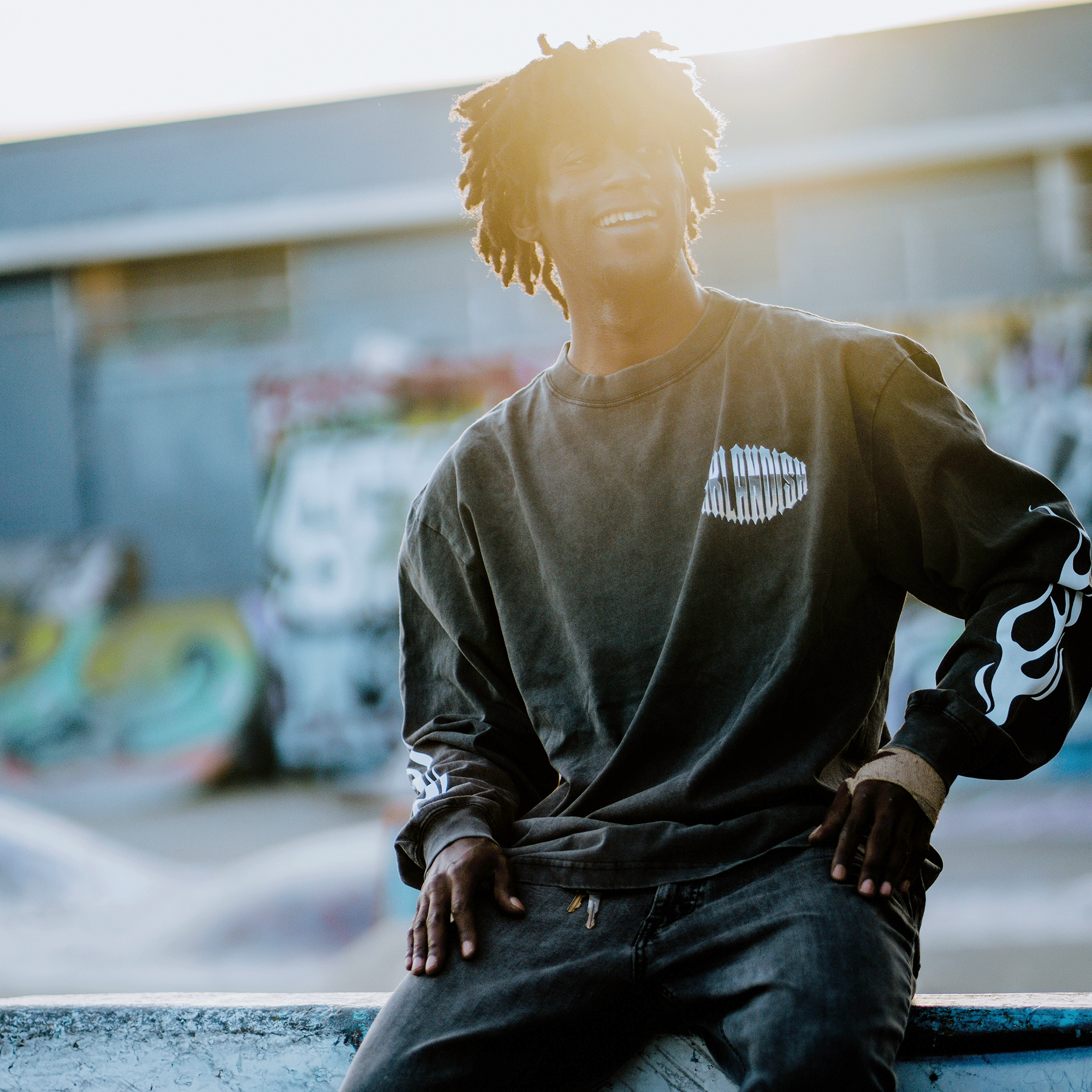 A person with curly hair sits on a concrete ledge in a skatepark, wearing the Chopper Heavy LS Tee by Oaklandish. Blurred graffiti decorates the walls behind them, while sunlight casts a bright halo effect, adding warmth to the scene.