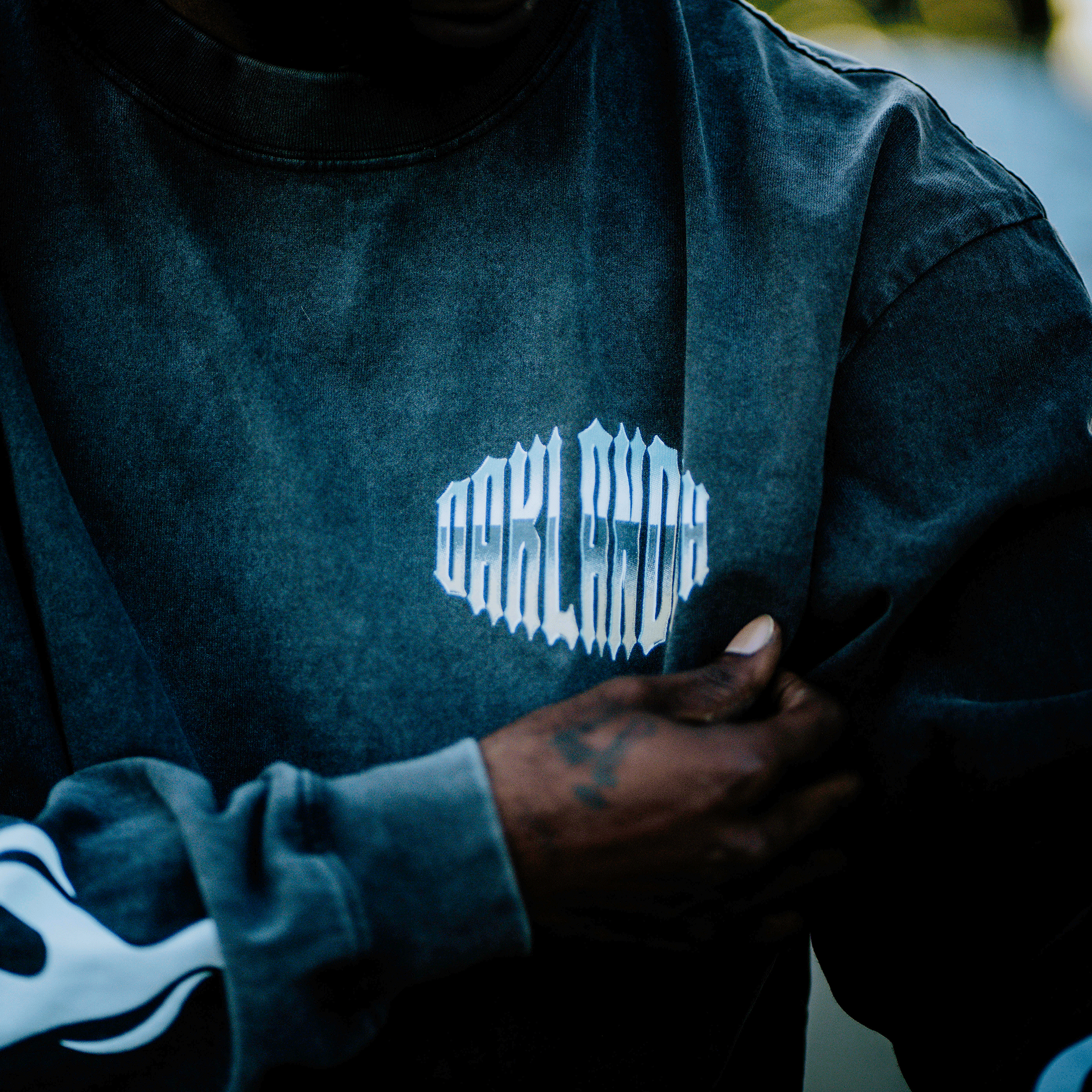 A person dons the vintage Chopper Heavy LS Tee by Oaklandish, a black long-sleeve with "OAKLAND" in white stylized letters and a white flame design on the sleeve. A hand tattoo stands out against the blurred backdrop, highlighting this 100% cotton piece's finer details.
