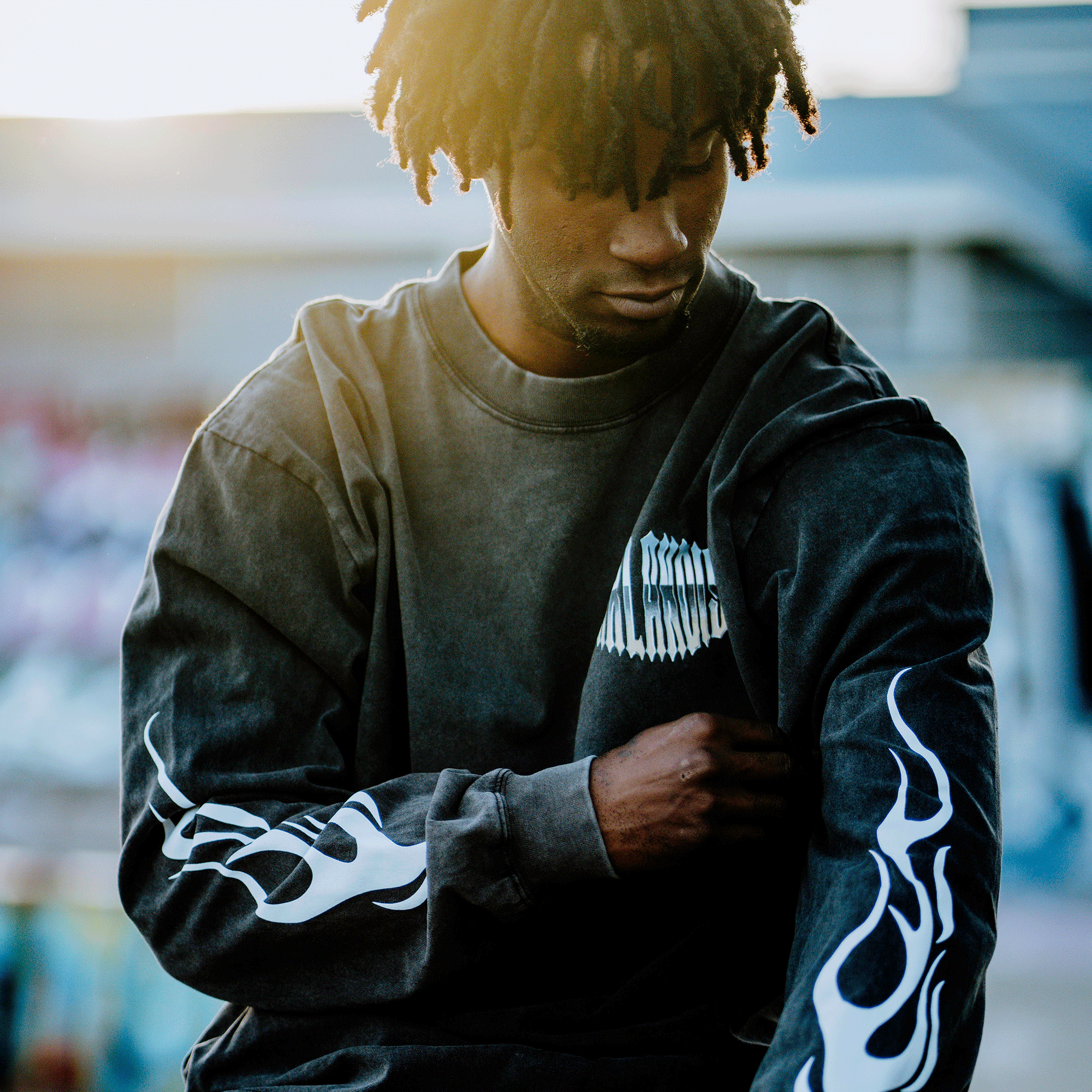 An individual with dreadlocks wears the Oaklandish Chopper Heavy LS Tee, a vintage black, long sleeve, 100% cotton shirt featuring white flame designs on the sleeves and text on the chest. They look downward in a sunlit, graffiti-blurred background, gently touching their sleeve.