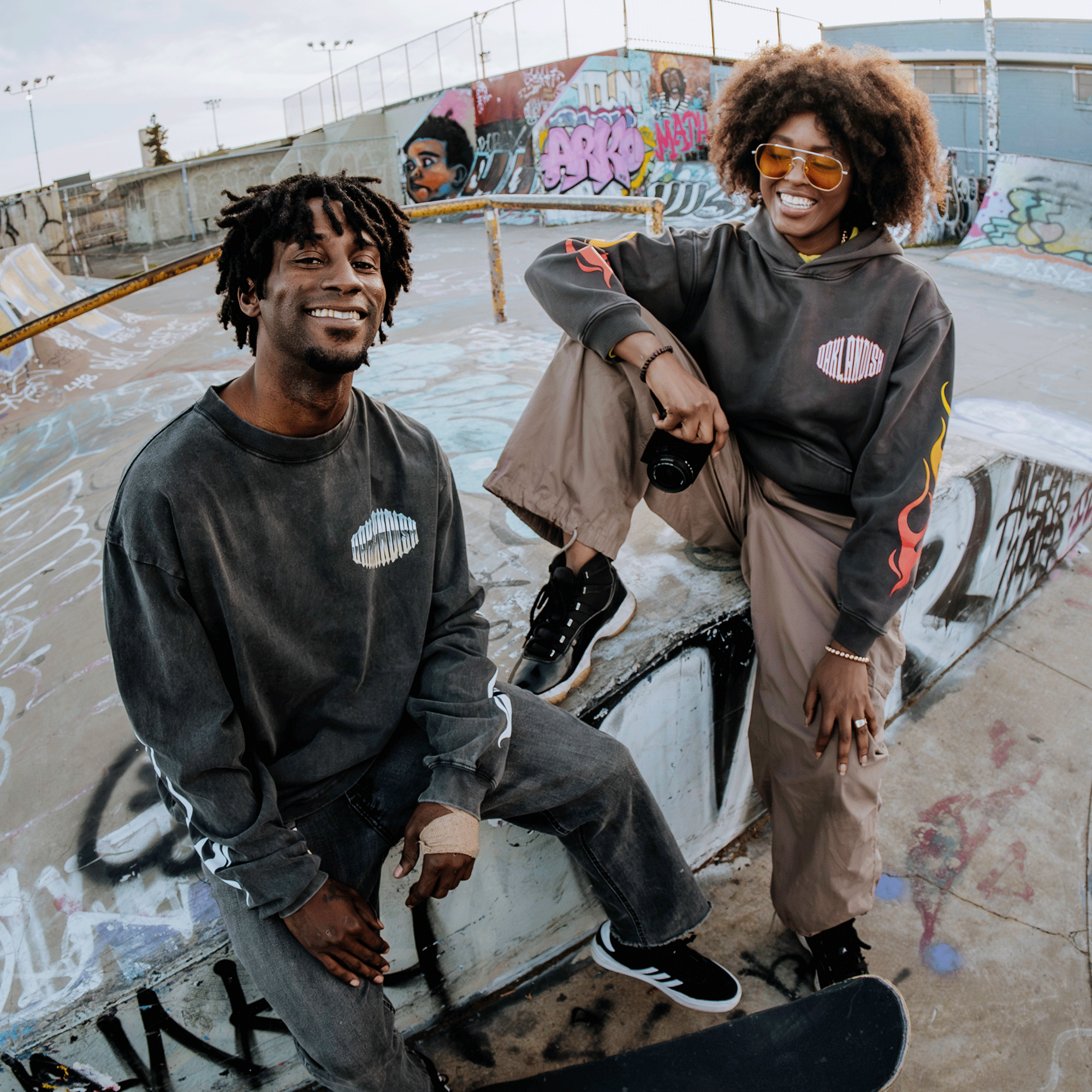 Two people in casual streetwear, featuring the Chopper Heavy Hoodie by Oaklandish, are on a graffiti-covered skate ramp. One has a skateboard, the other wields a camera. Both smile amidst vibrant urban art and clear skies, capturing a relaxed and creative vibe.