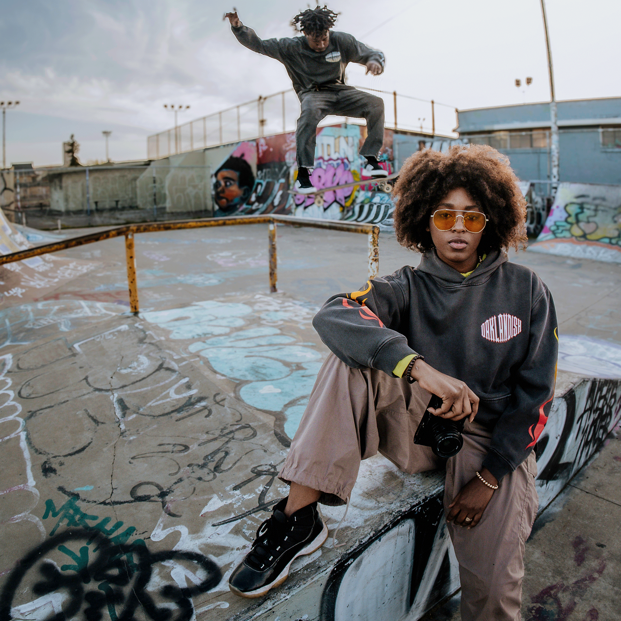 A young woman in an oversized Chopper Heavy Hoodie by Oaklandish and drop-shoulder sunglasses sits on a graffiti-covered skatepark ramp, clutching her camera. In the background, someone catches air on a skateboard against an urban backdrop of cloudy skies and buildings.