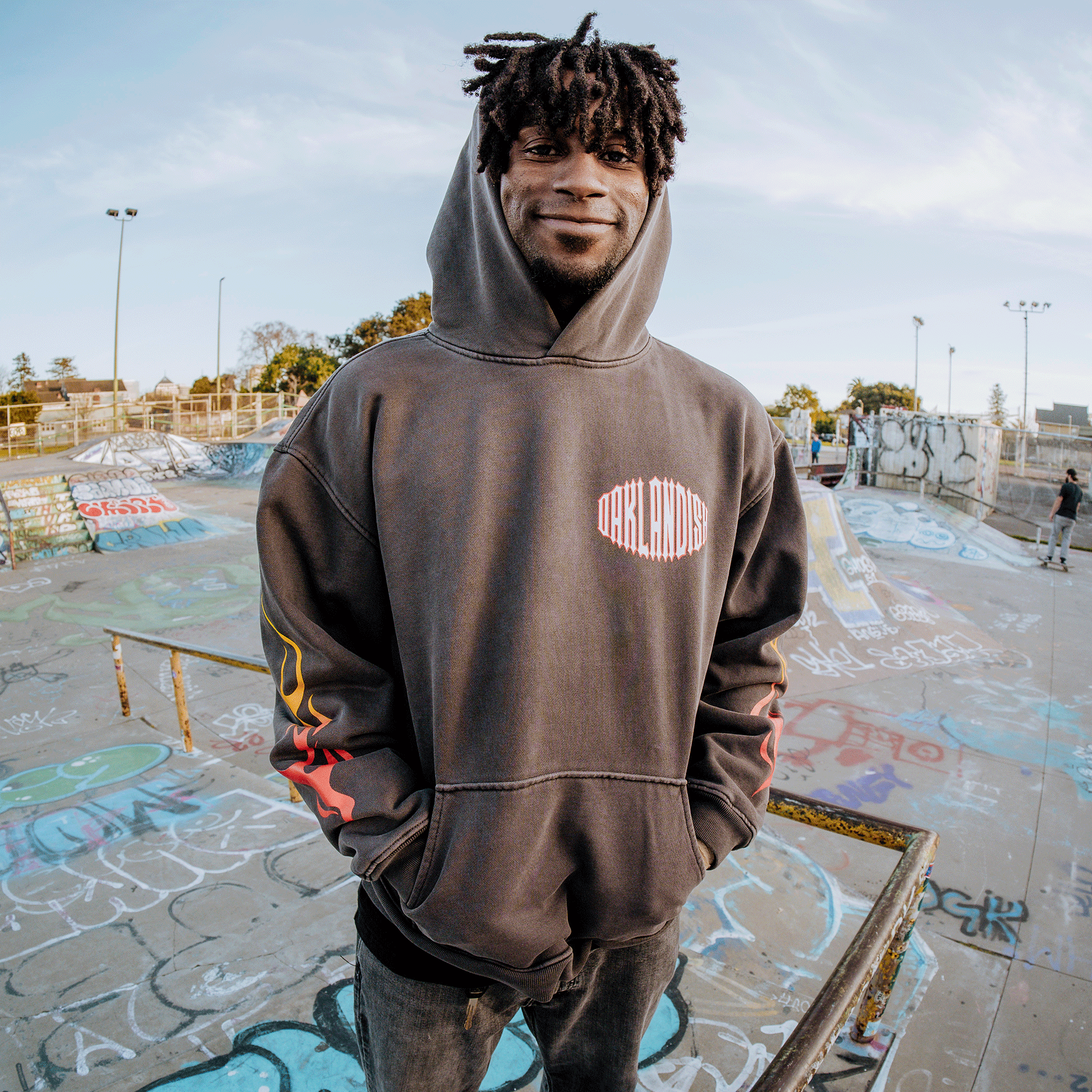 A person confidently stands in a graffiti-covered skate park wearing the Chopper Heavy hoodie by Oaklandish. This oversized drop-shoulder pullover, made from 100% cotton fleece, drapes over their dreadlocks as they rest their hands in the pockets, with ramps and a clear blue sky behind them.