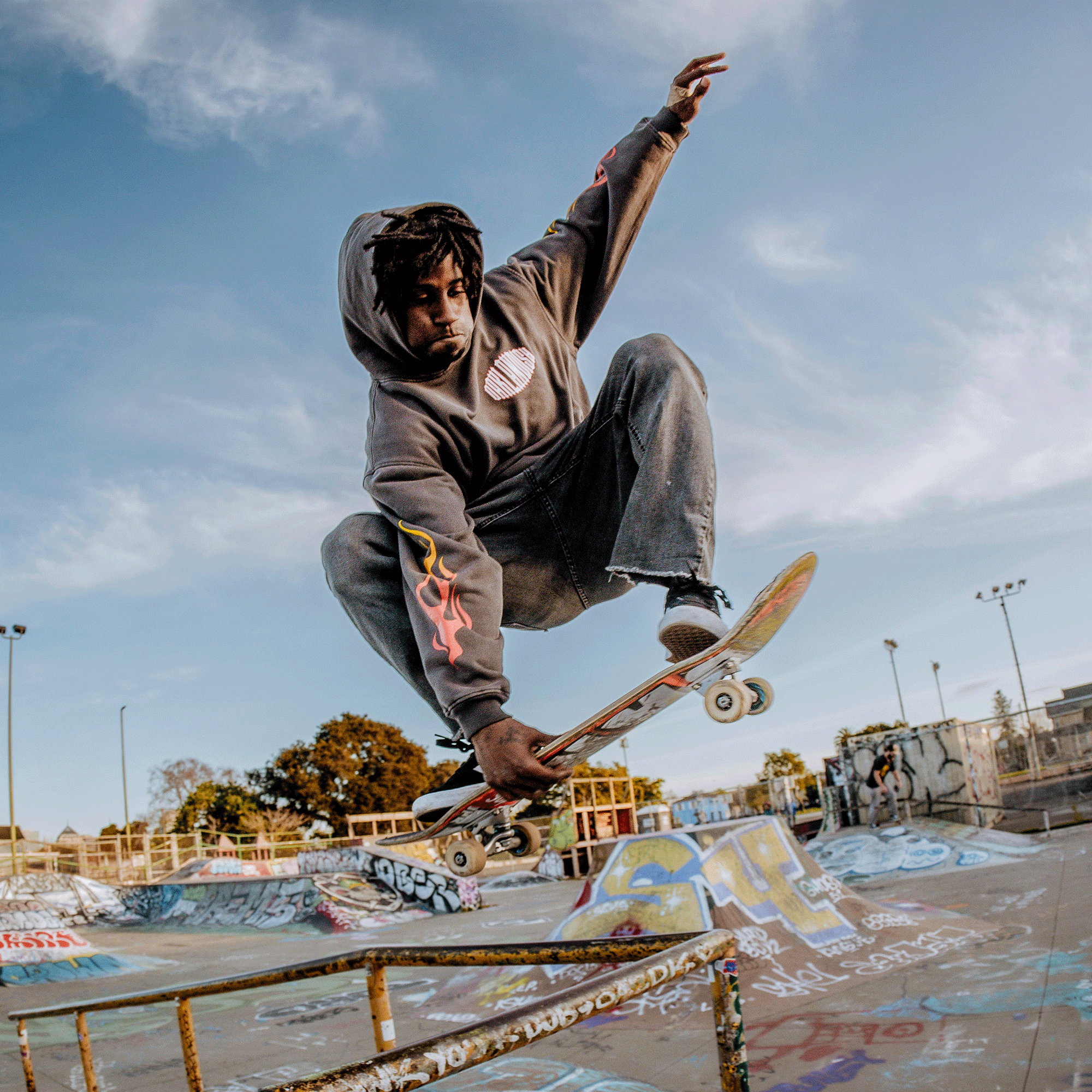 A skateboarder in an oversized Chopper Heavy Hoodie by Oaklandish and jeans performs an aerial trick above a rail at a vibrant, graffiti-covered skatepark. The sky is clear with scattered clouds, and trees are visible in the background, capturing dynamic motion and focus.