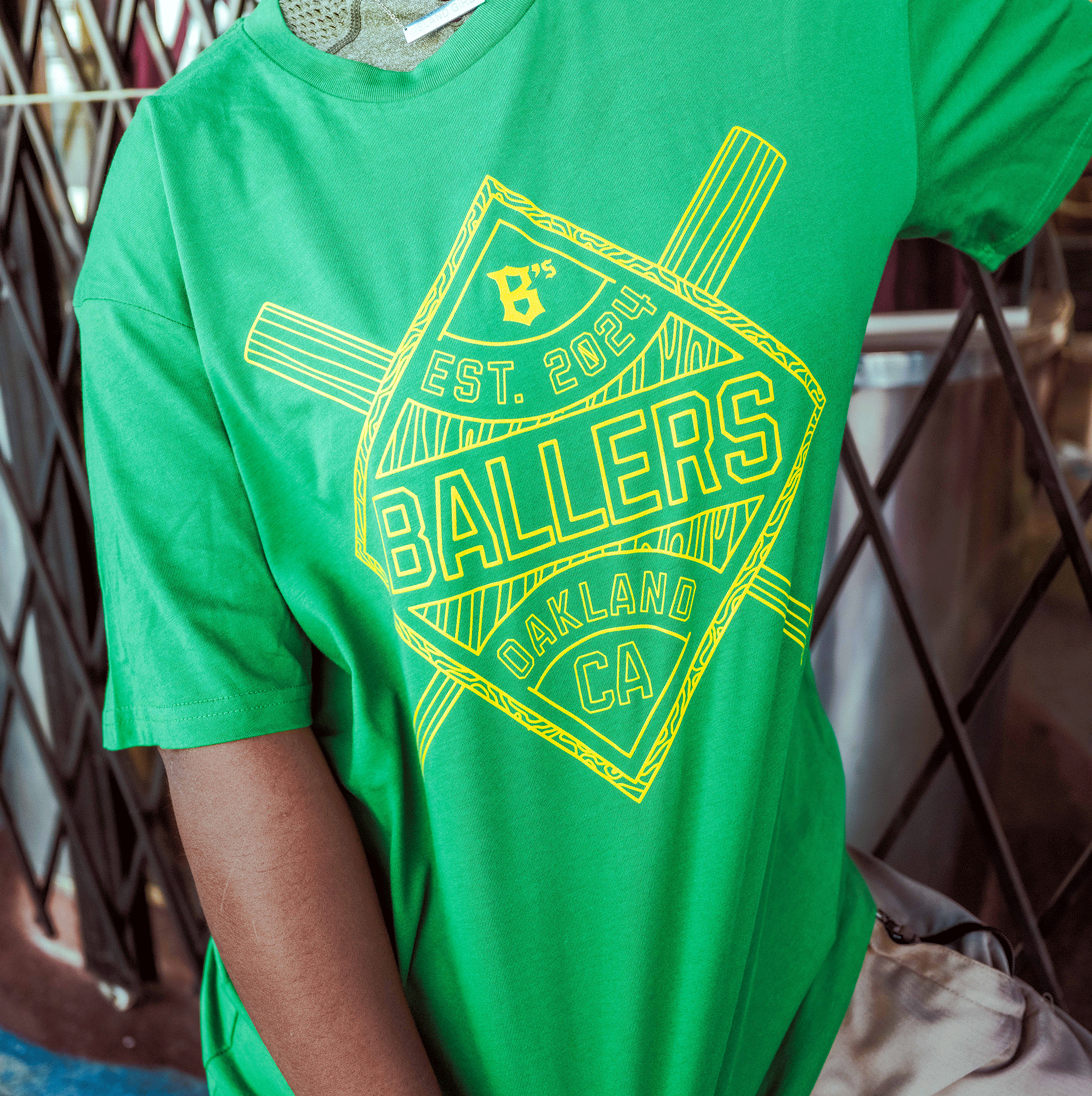 A person wears the Oakland Ballers Classic Diamond Tee in green, featuring yellow graphics—“BALLERS,” “OAKLAND CA,” “EST. 2024,” geometric line art, and a small baseball diamond—with a metal gate and outdoor scenery in the background.