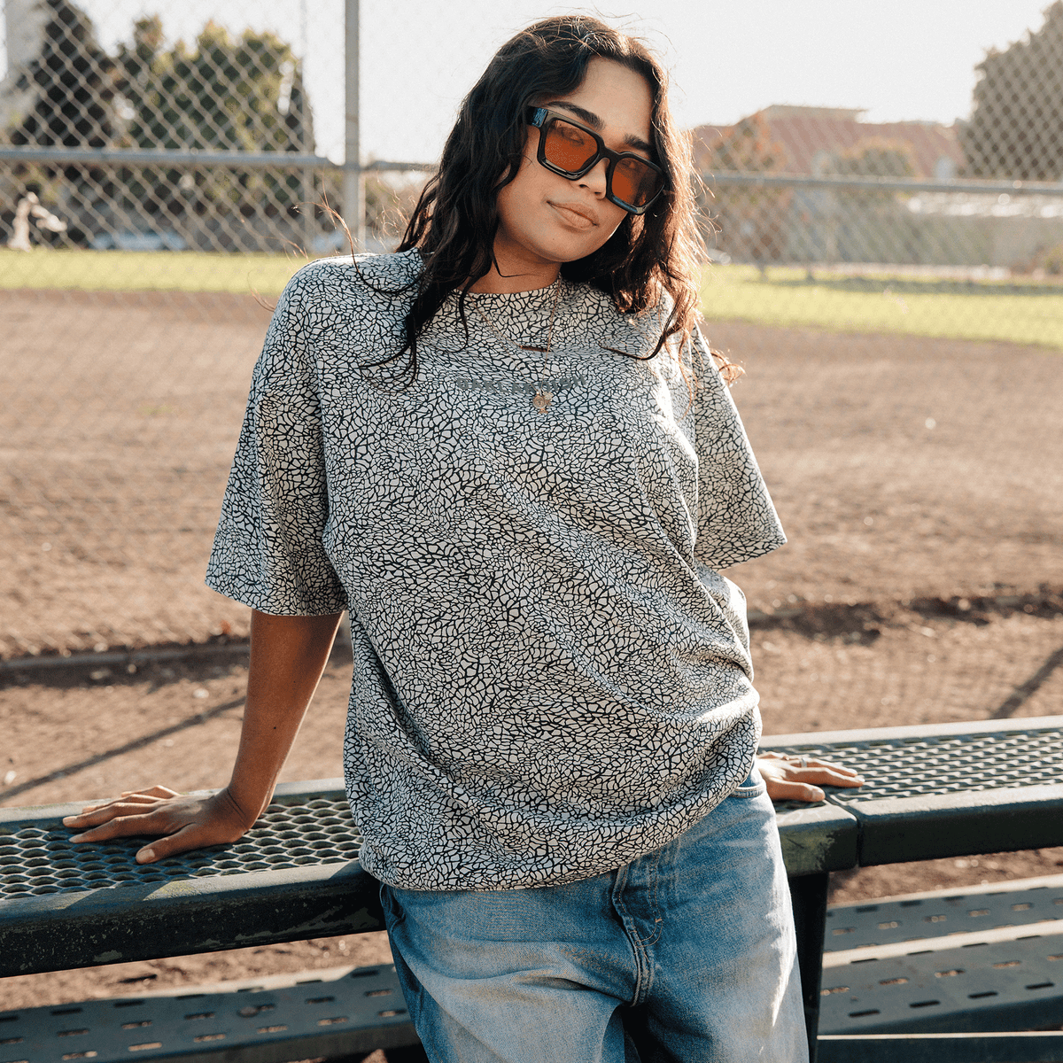 A person with long dark hair and tinted sunglasses wears an Oaklandish Classic Logo Tee and loose-fitting blue jeans while leaning back against a green metal bench on a dirt baseball field near a chain-link fence.