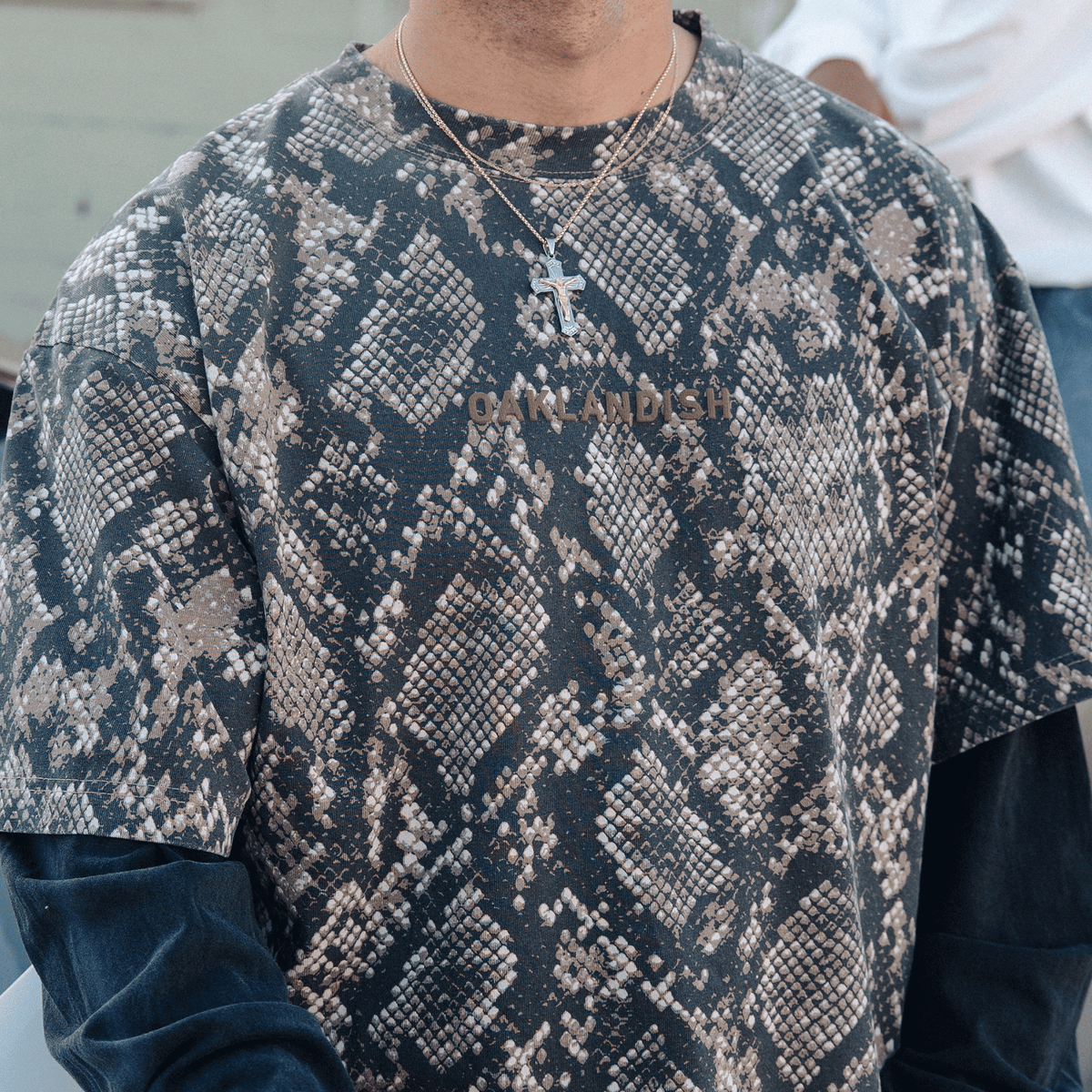 A person wears a vintage black long-sleeved shirt under an Oaklandish Classic Logo LS Tee featuring a grey and brown snakeskin pattern with “OAKLANDISH” printed across the chest, and a silver crucifix hanging on a thin chain around their neck.
