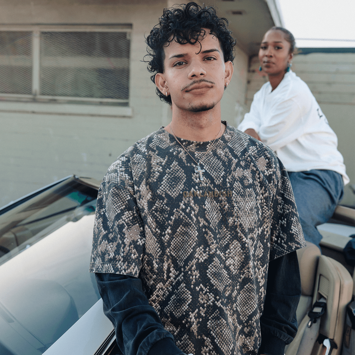A person with dark curly hair wears a short-sleeved, snakeskin-patterned shirt layered over a vintage black Oaklandish Classic Logo LS Tee, standing in front of a parked car with a second person leaning against it in the background.