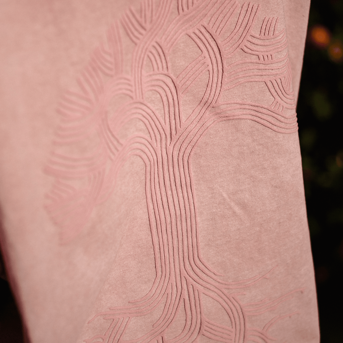 A close-up of a dusty pink Oaklandish Classic Logo Tee featuring a tonal, embossed oak tree design with a puff print pattern of sweeping branches and roots that blend into the fabric’s soft hue.