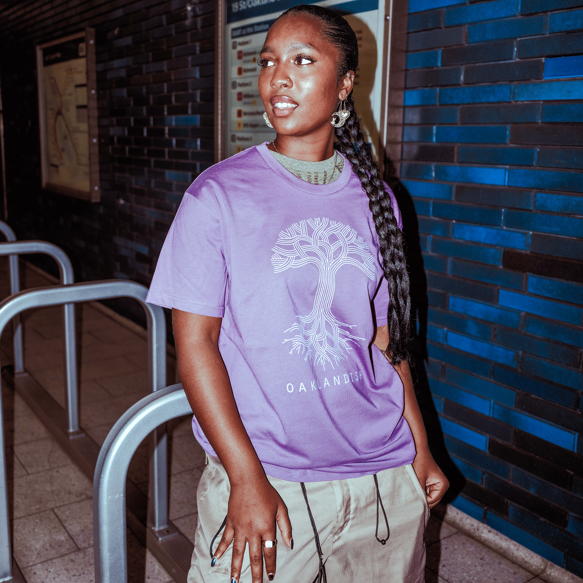 A person with long braids stands in a subway station, wearing the Oaklandish Classic Logo Tee by Oaklandish, khaki pants, and silver earrings. Blue tile walls and bike racks can be seen in the background.