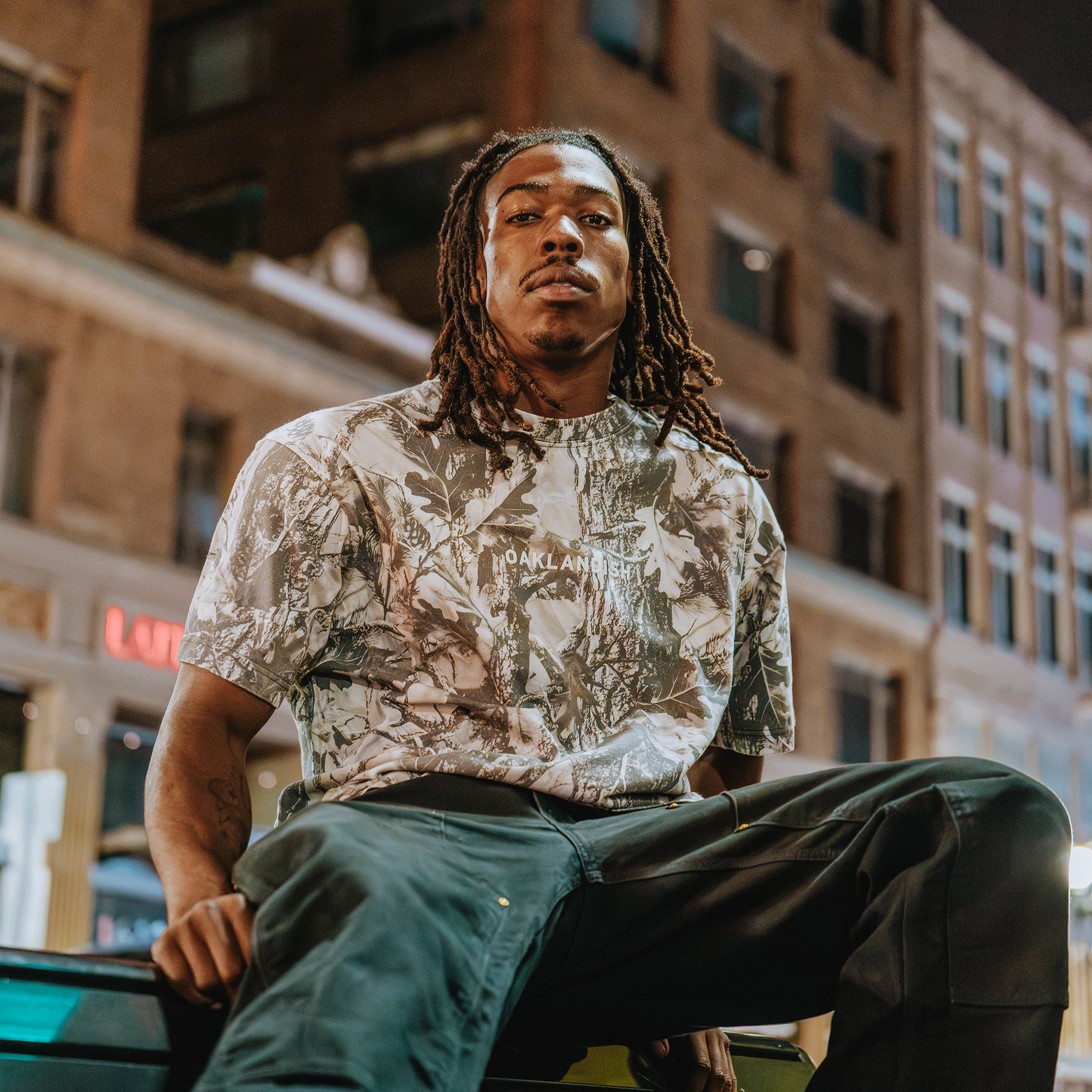 A young man with long dreadlocks sits on a ledge at night in the city, wearing the Oaklandish Classic Logo Tee by Oaklandish—crafted from heavyweight 100% cotton—and dark pants. Neon lights and tall buildings add to the urban vibe.