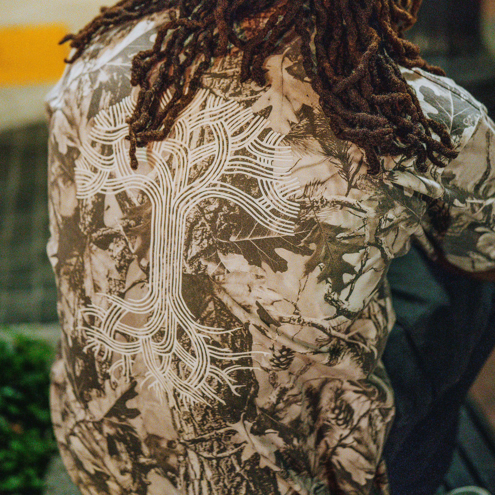 A person with long, thick dreadlocks sits facing away, wearing the Oaklandish Classic Logo Tee by Oaklandish. The background features greenery and a blurred wall with yellow stripes.