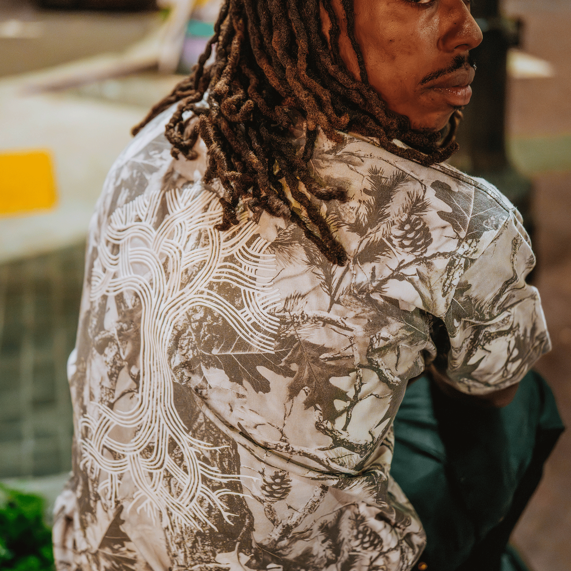 A person with long, brown dreadlocks sits turned away from the camera, glancing back. They're wearing an Oaklandish Classic Logo Tee—a heavyweight 100% cotton shirt—against a blurred urban background.