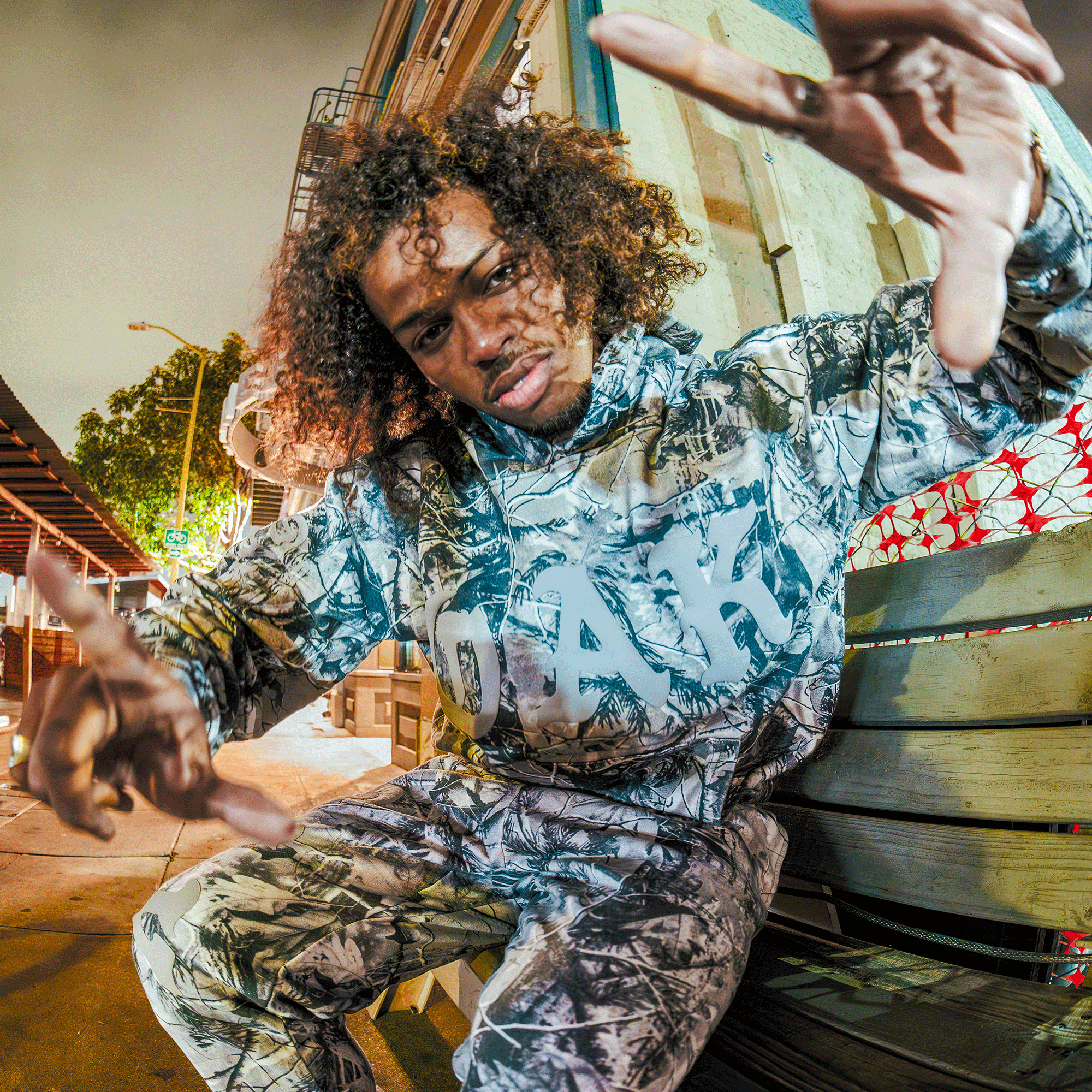 Wearing a Grey Wood Camo hoodie and Oaklandish Cork Pant, a curly-haired person poses energetically on a wooden bench along a city sidewalk at night, surrounded by colorful lights and urban scenery.