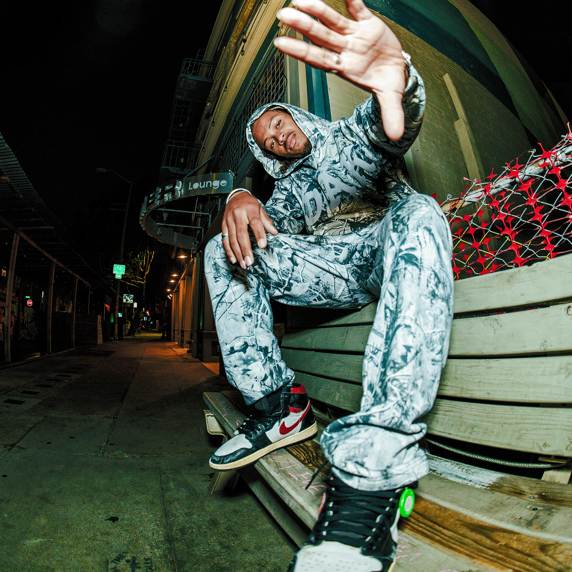A person wearing the Oaklandish Cork Pant and a matching Grey Wood Camo cotton hoodie sits on a wooden bench at night, reaching toward the camera. They have black, white, and red sneakers; nearby are a lounge sign and construction barriers.